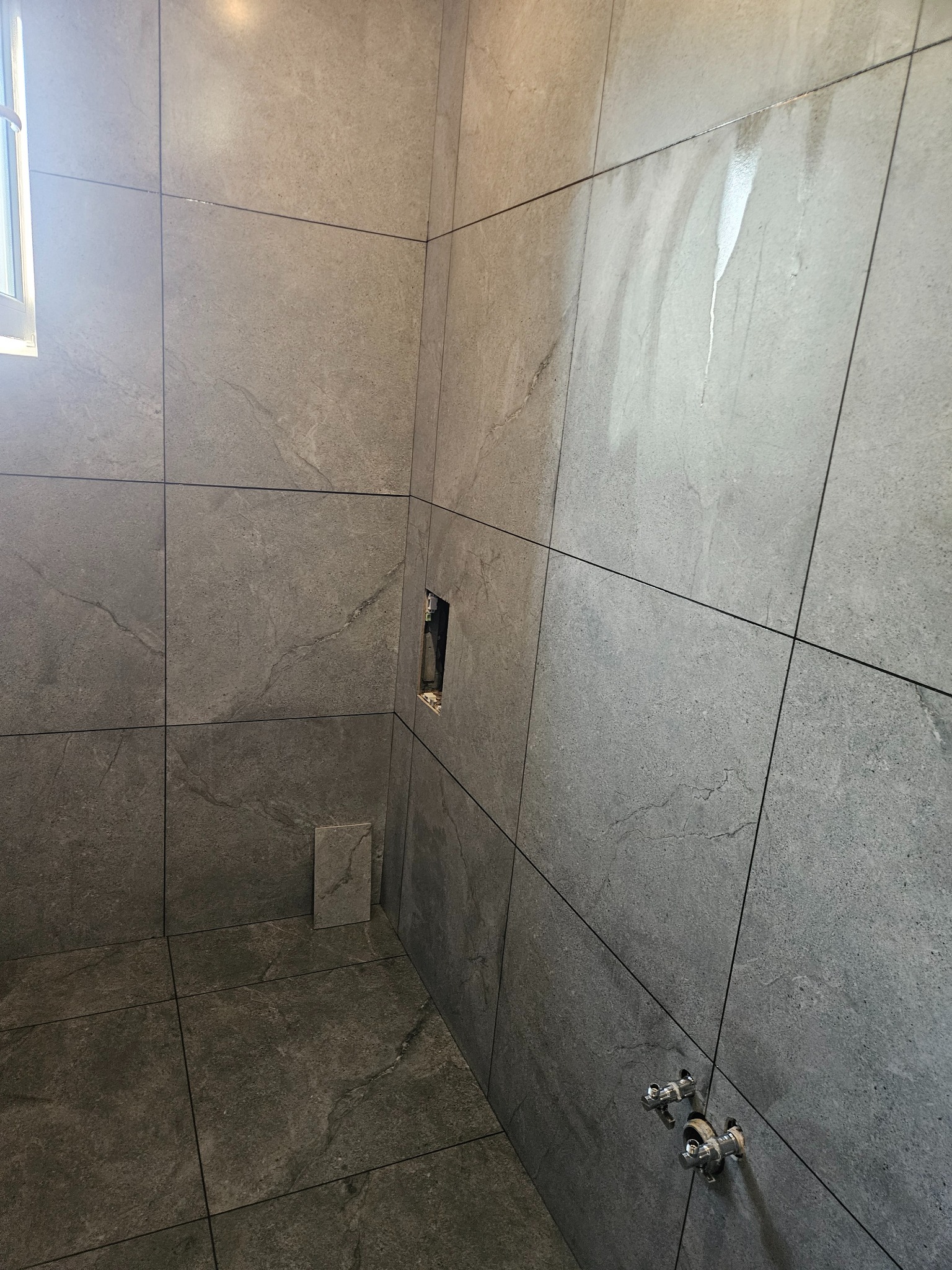 Wykończona łazienka z szarymi, wielkoformatowymi płytkami. Widoczne przyłącza wodne i otwór na armaturę. Minimalistyczny design z oknem w górnym lewym rogu.