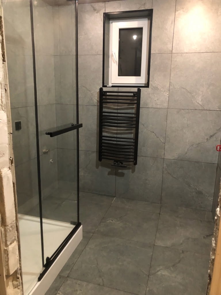 Wykończona łazienka z kabiną prysznicową typu walk-in, grzejnikiem drabinkowym i oknem. Szare płytki imitujące kamień na ścianach i podłodze. Minimalistyczny design.