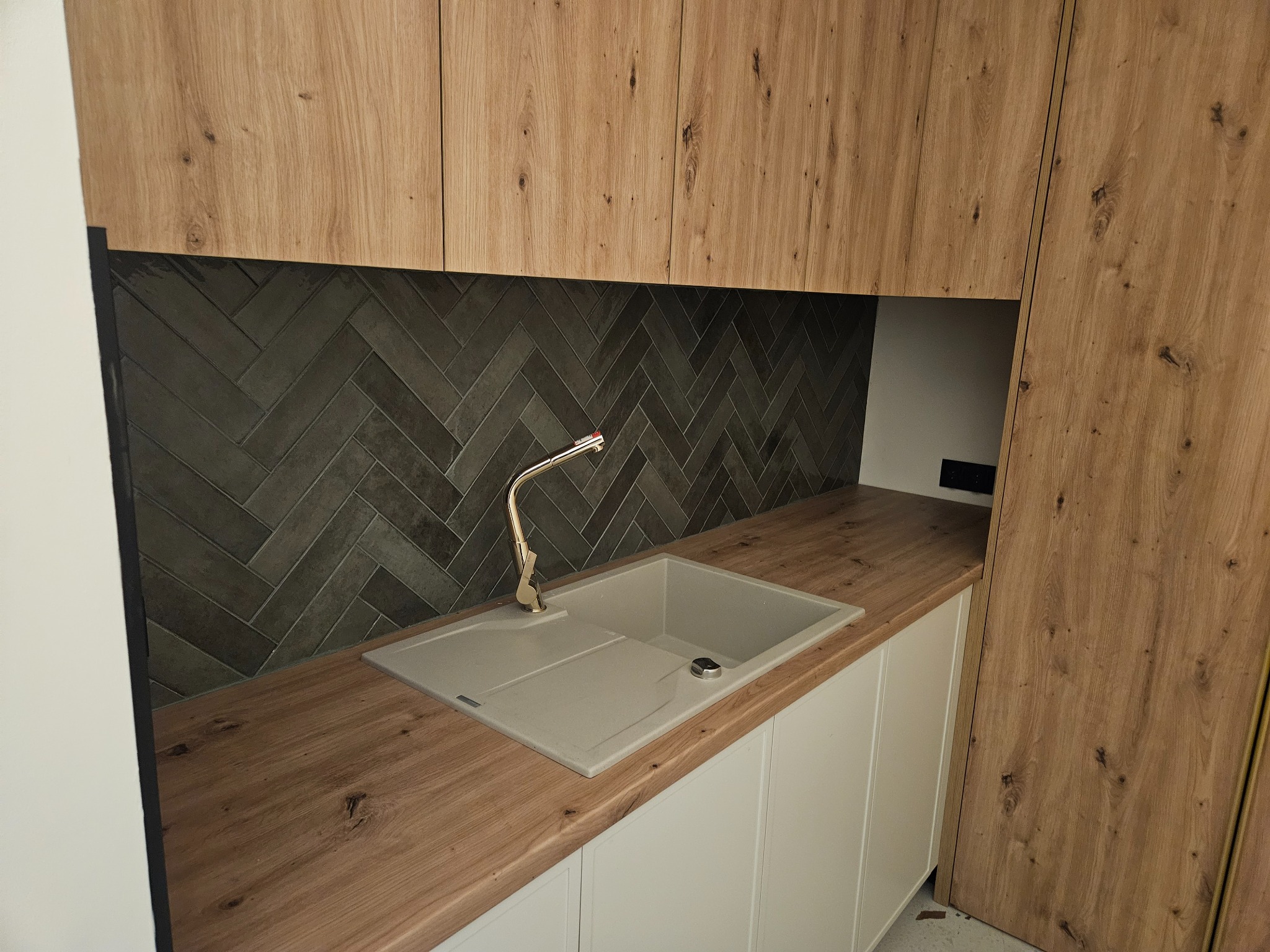 Nowoczesna kuchnia z drewnianymi szafkami i szarą, ułożoną w jodełkę ścianą nad blatem. Jasny zlew z baterią. Minimalistyczny design.