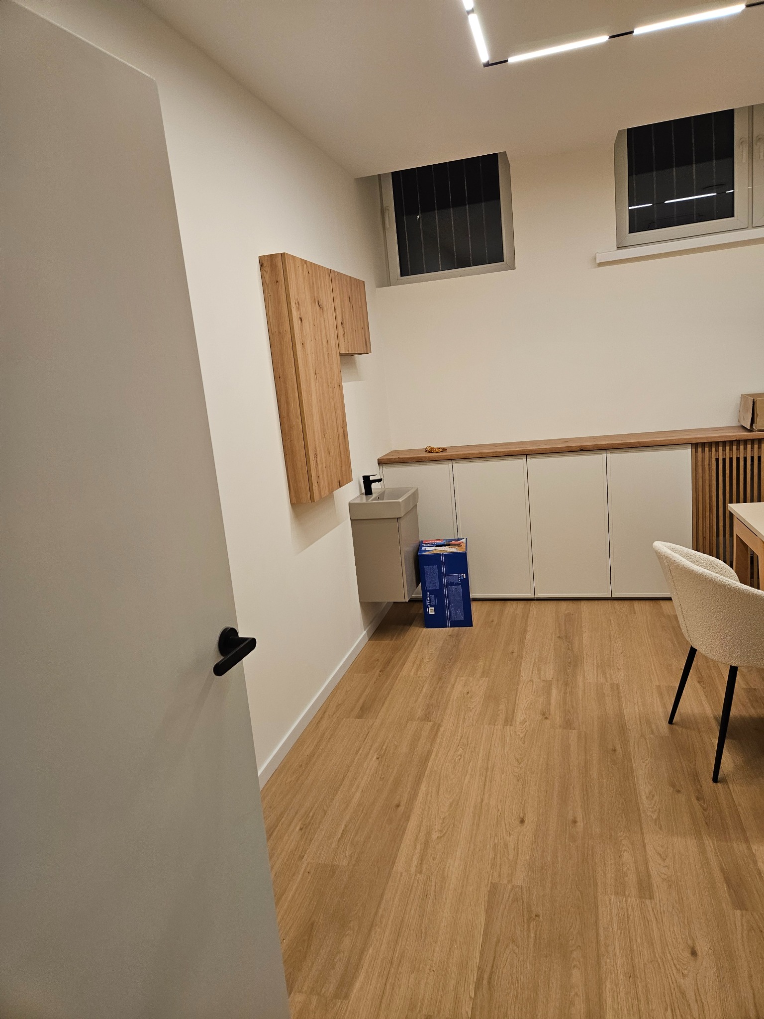 Nowoczesna łazienka z drewnianymi akcentami: szafka nad umywalką, blat z szafkami, podłoga z paneli. Widoczne okna z kratami i minimalistyczne oświetlenie.