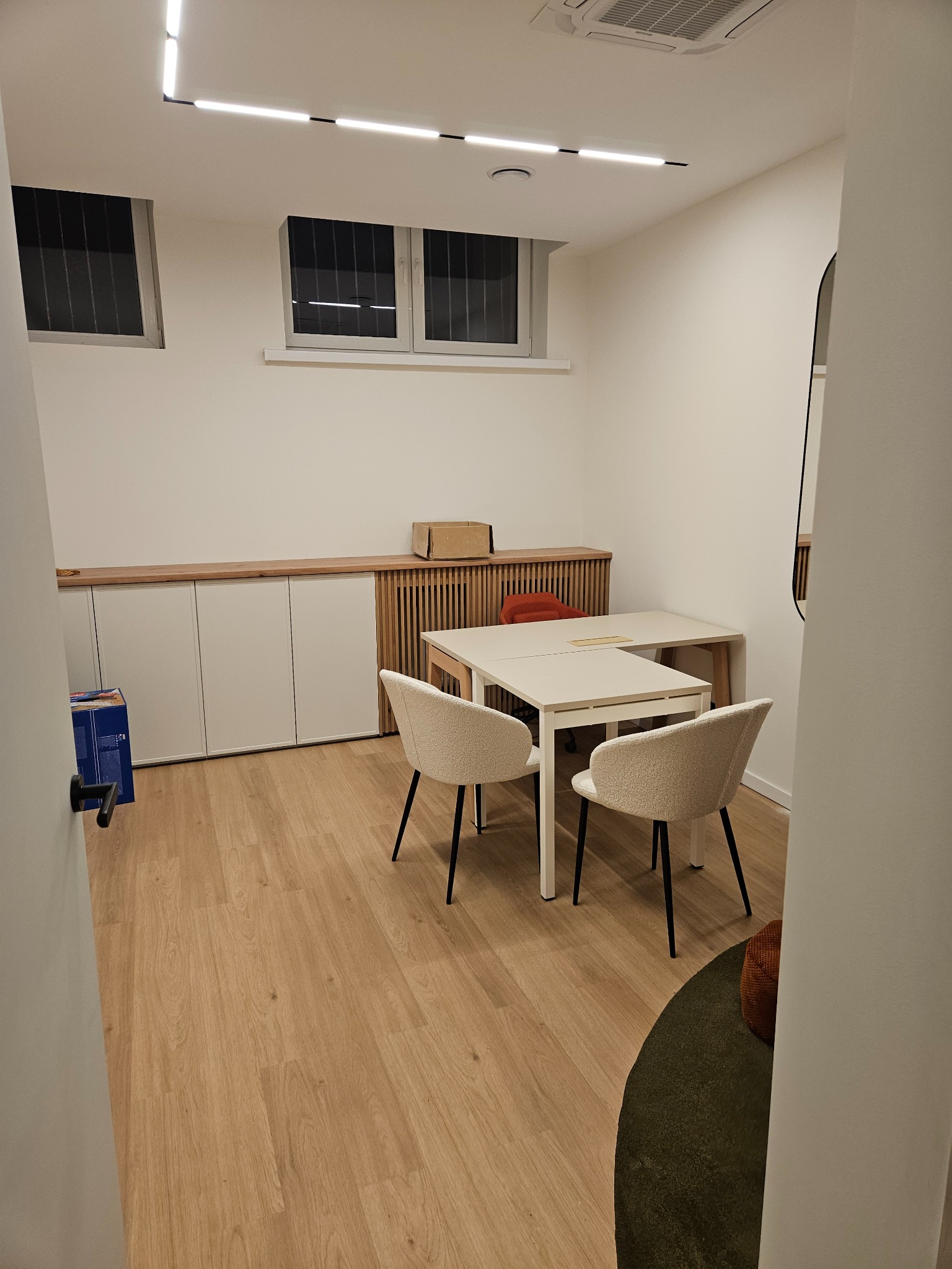 Jasne biuro z drewnianą podłogą, białymi ścianami i meblami. Widoczne biurko, krzesła i szafki w zabudowie. Minimalistyczny design z oświetleniem LED.