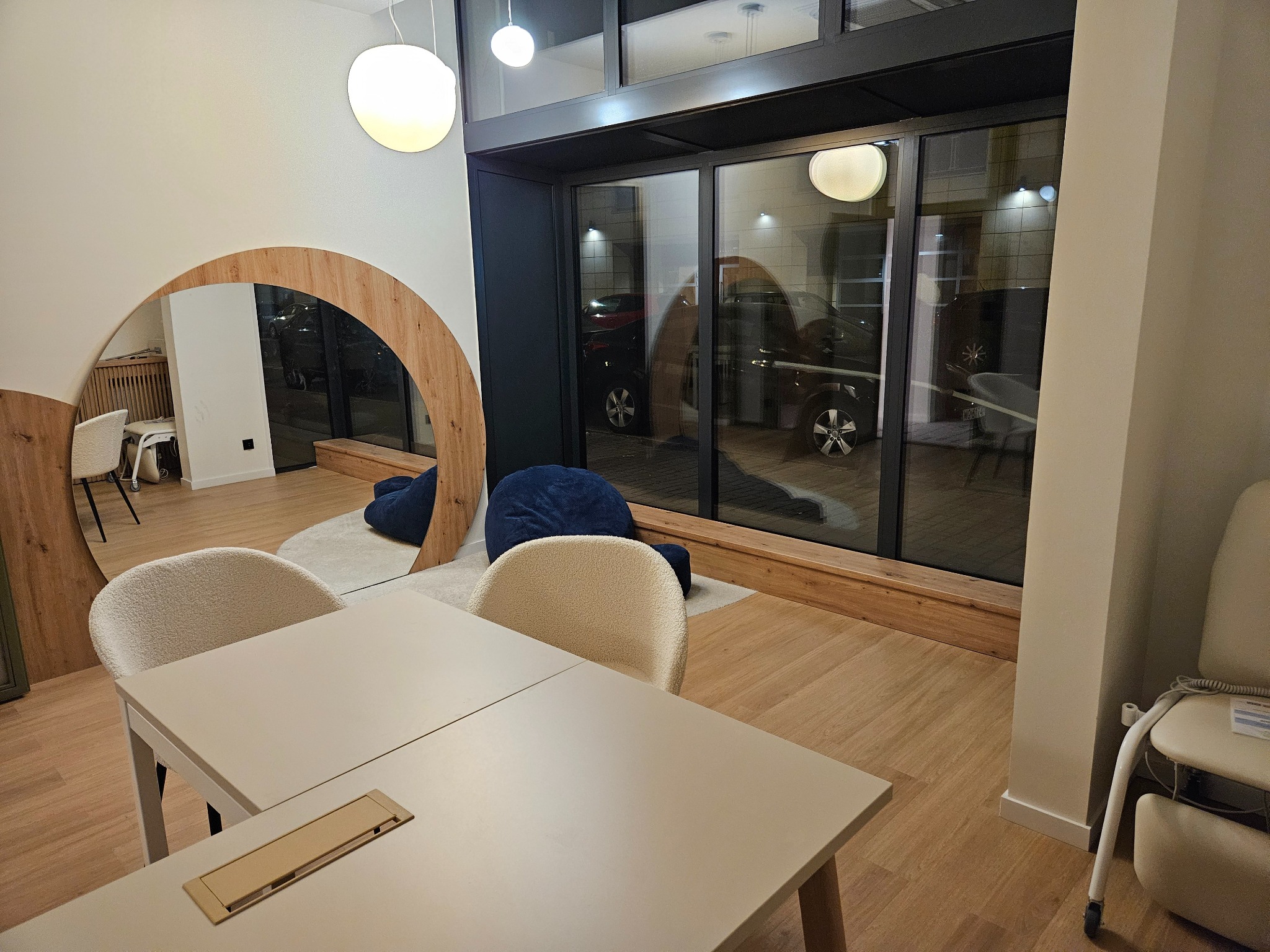 Nowoczesne biuro z drewnianym lustrem w kształcie łuku, beżowymi krzesłami i stołem. Za oknem widoczny parking. Minimalistyczny design wnętrza.