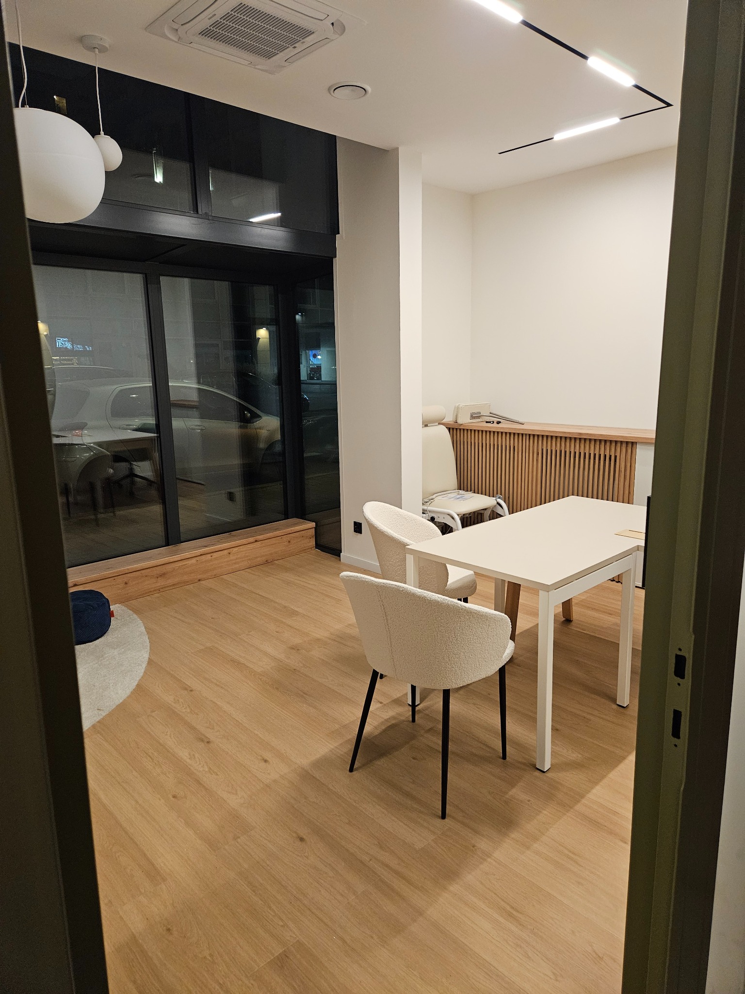 Jasne, minimalistyczne wnętrze biurowe z drewnianą podłogą, białym stołem i krzesłami. Widoczne duże okno z widokiem na ulicę i nowoczesne oświetlenie sufitowe.