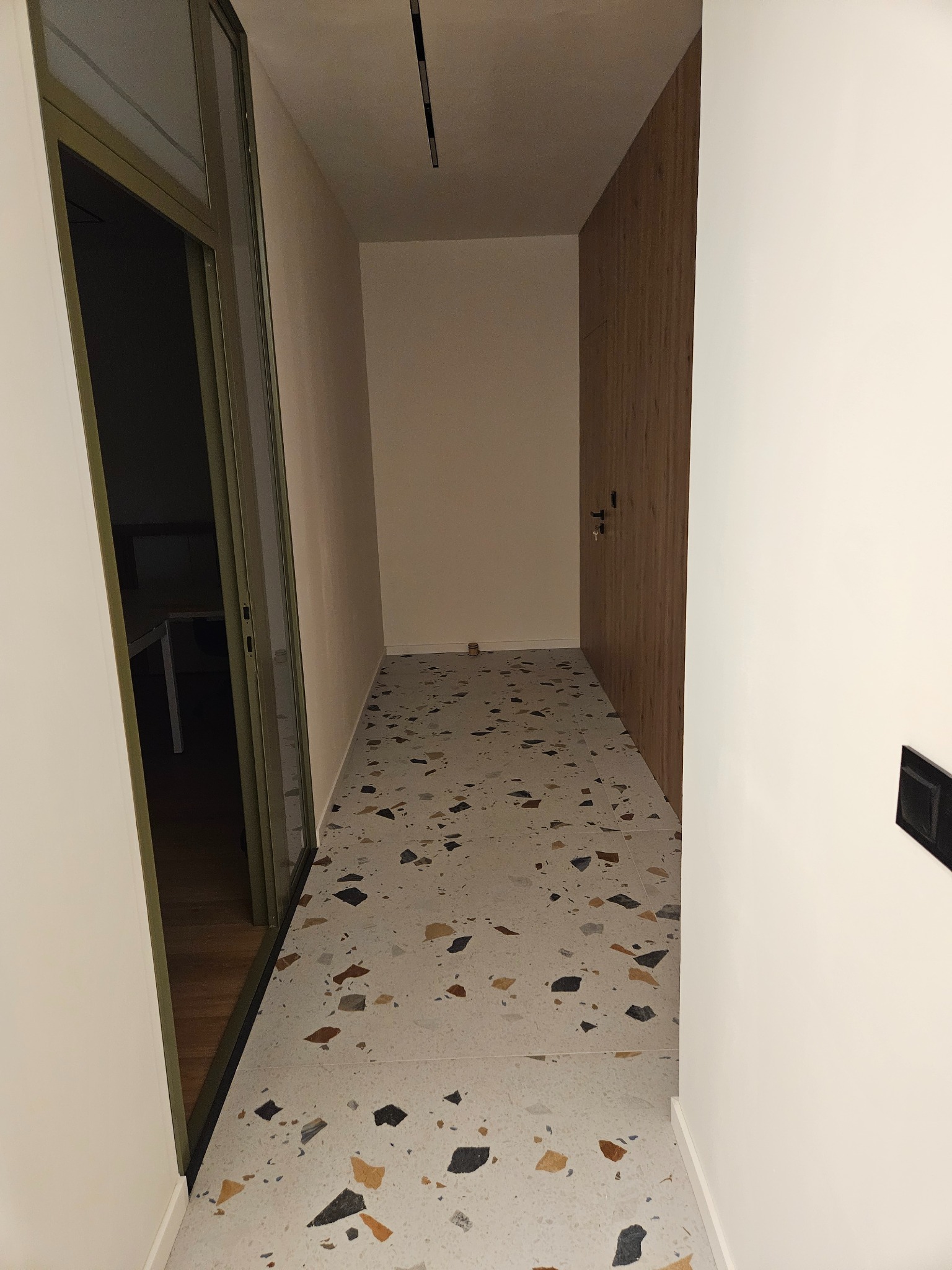 Korytarz z terazzo na podłodze, widok z perspektywy wejścia. Ściany w odcieniach bieli i beżu, drewniane drzwi po prawej. Widoczny fragment ramy drzwiowej po lewej.