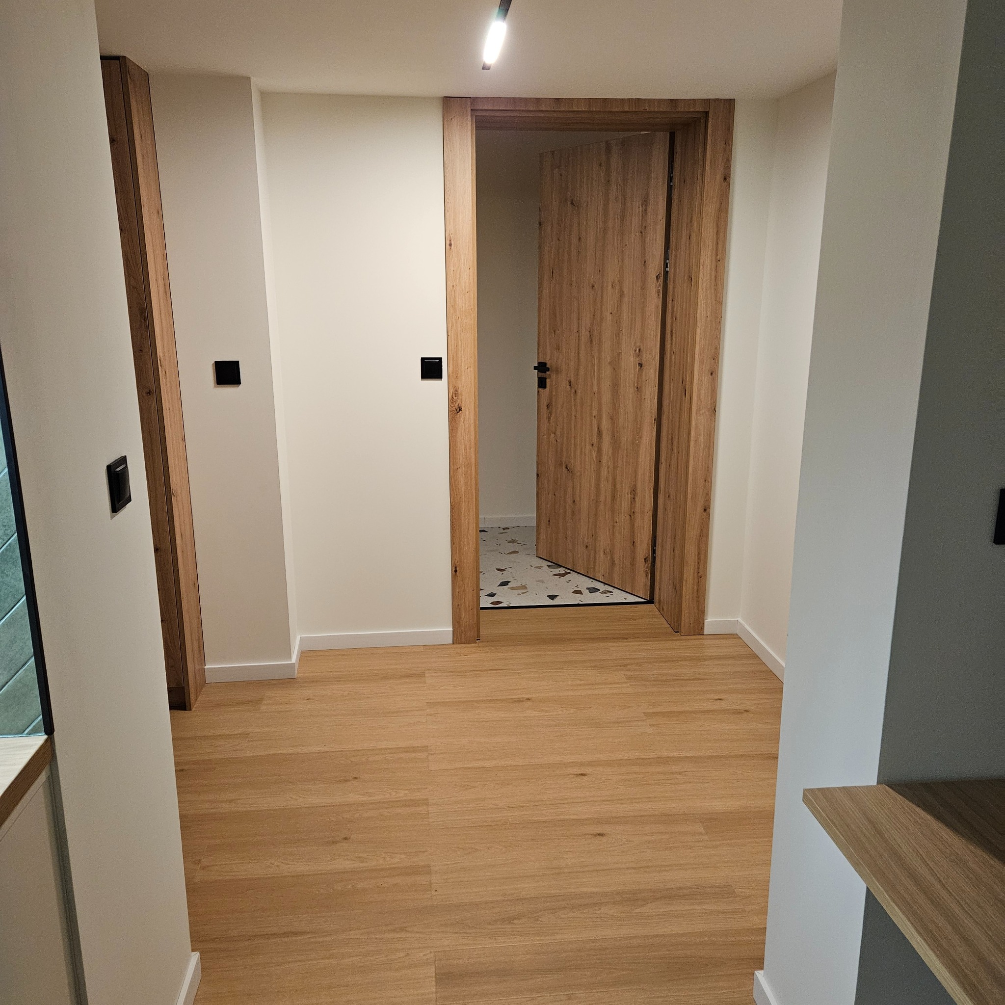 Jasne panele podłogowe w korytarzu z drewnianą ościeżnicą i drzwiami. W tle widoczna podłoga z mozaiką. Minimalistyczne wykończenie wnętrza.