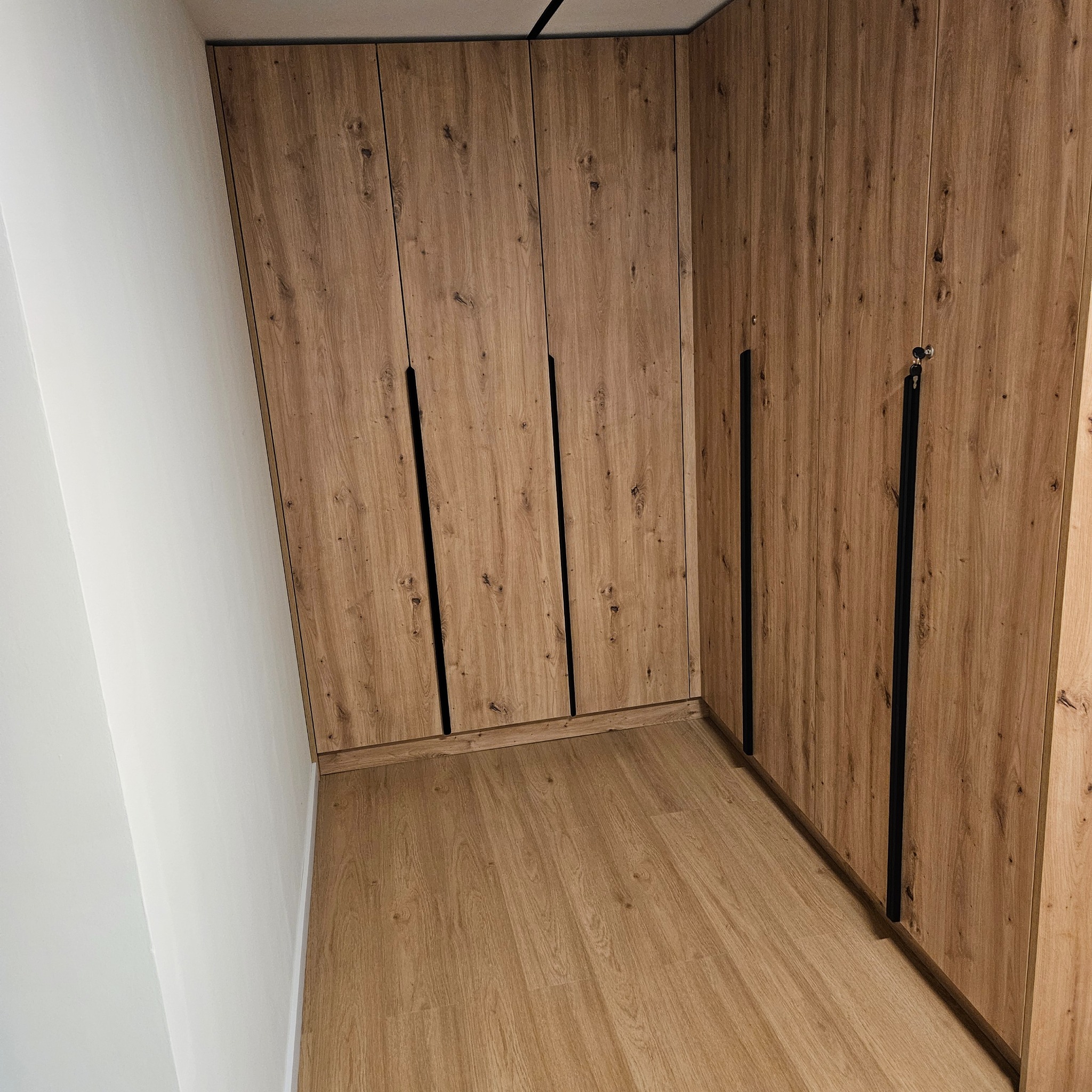 Wnętrze z zabudową szafy w odcieniach drewna, z czarnymi, pionowymi uchwytami, dopasowaną podłogą i białymi ścianami. Widok z perspektywy podłogi.