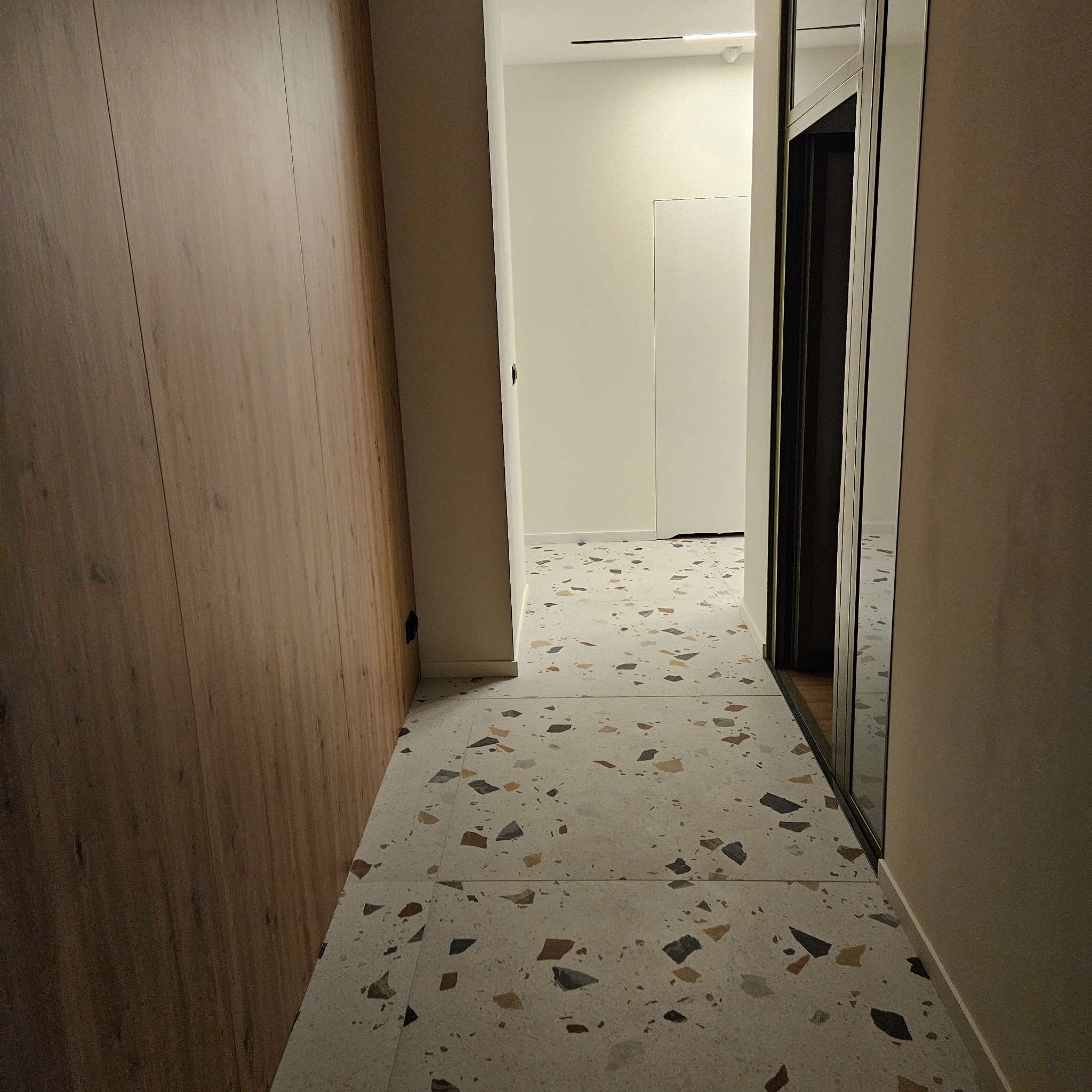 Korytarz z drewnianą zabudową i podłogą w stylu lastryko. Widoczne drzwi przesuwne i oświetlenie sufitowe. Minimalistyczny design wnętrza.