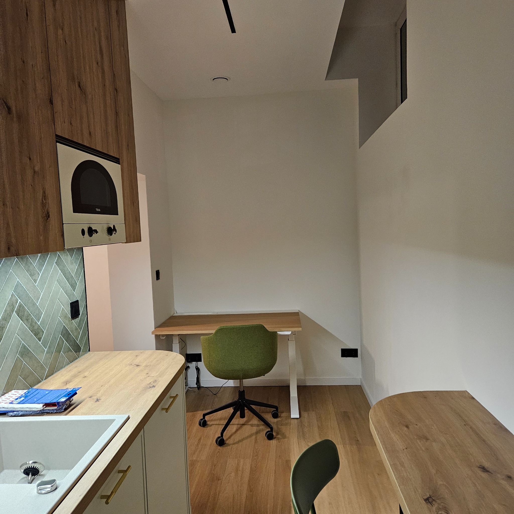 Nowoczesna aranżacja wnętrza: drewniane szafki kuchenne z piekarnikiem, zielona ściana z płytek w jodełkę, biurko z krzesłem i nietypowe okno w górnej części ściany.