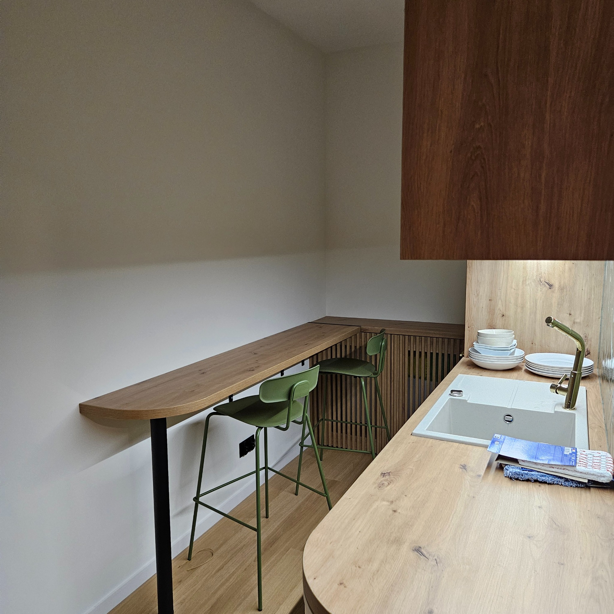 Nowoczesna kuchnia z blatem z jasnego drewna, zielonymi hokerami i białym zlewem. Uwagę przykuwa minimalistyczny design i ciepłe oświetlenie.