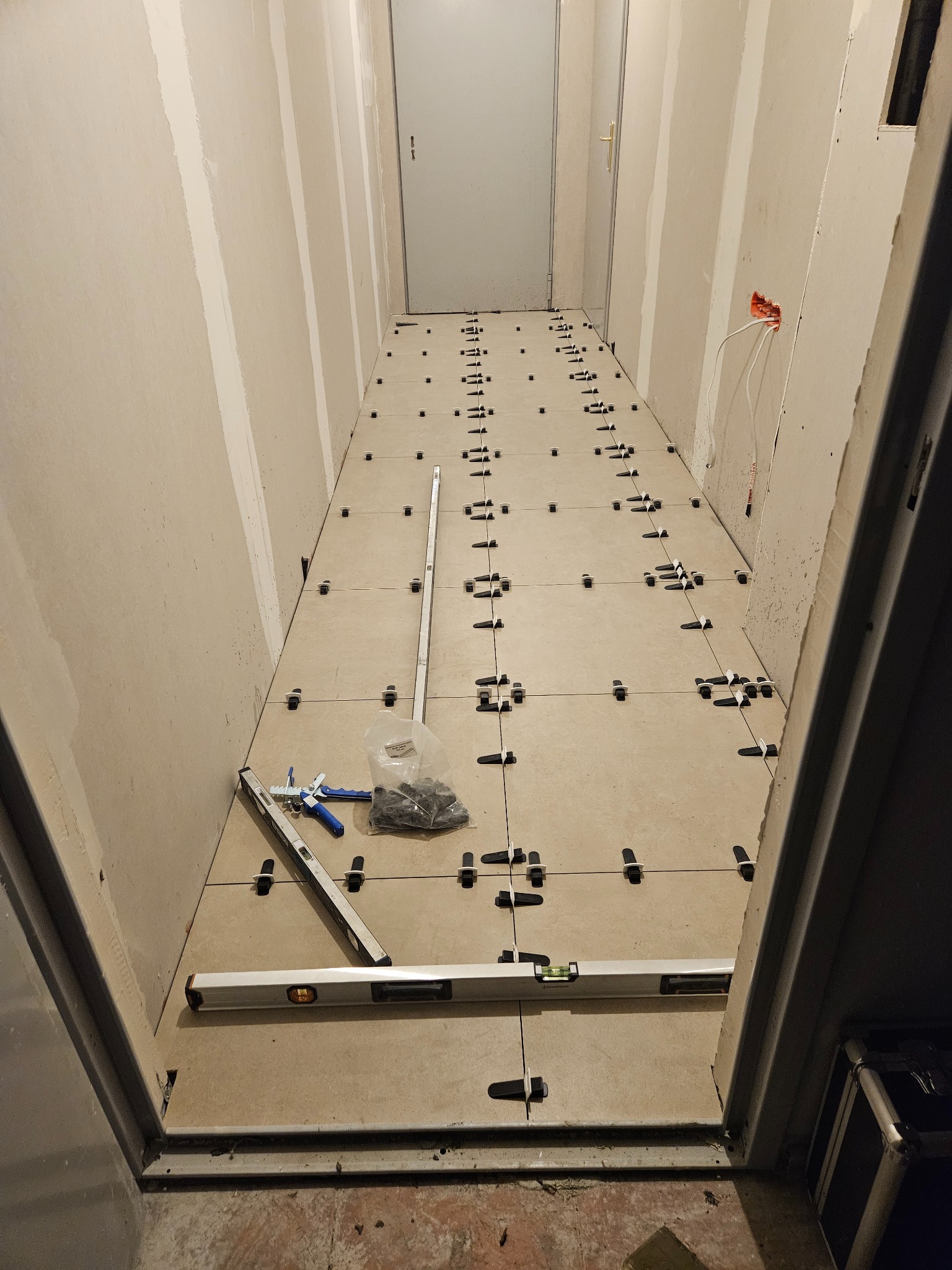 Układanie beżowych płytek podłogowych w wąskim korytarzu. Widoczne poziomice, paczka z klinami i szczypce do poziomowania. Ściany przygotowane pod malowanie.