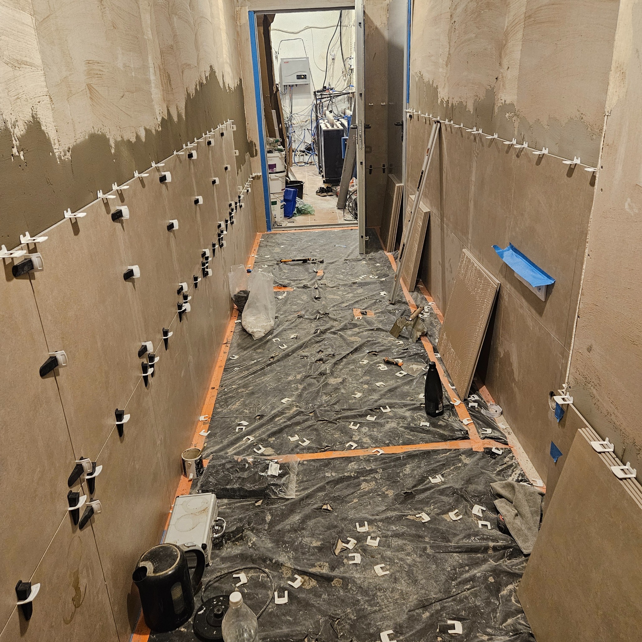 Korytarz w trakcie prac glazurniczych: częściowo wyłożone płytki na ścianach z użyciem klipsów poziomujących, podłoga zabezpieczona folią. Widoczne narzędzia i materiały.