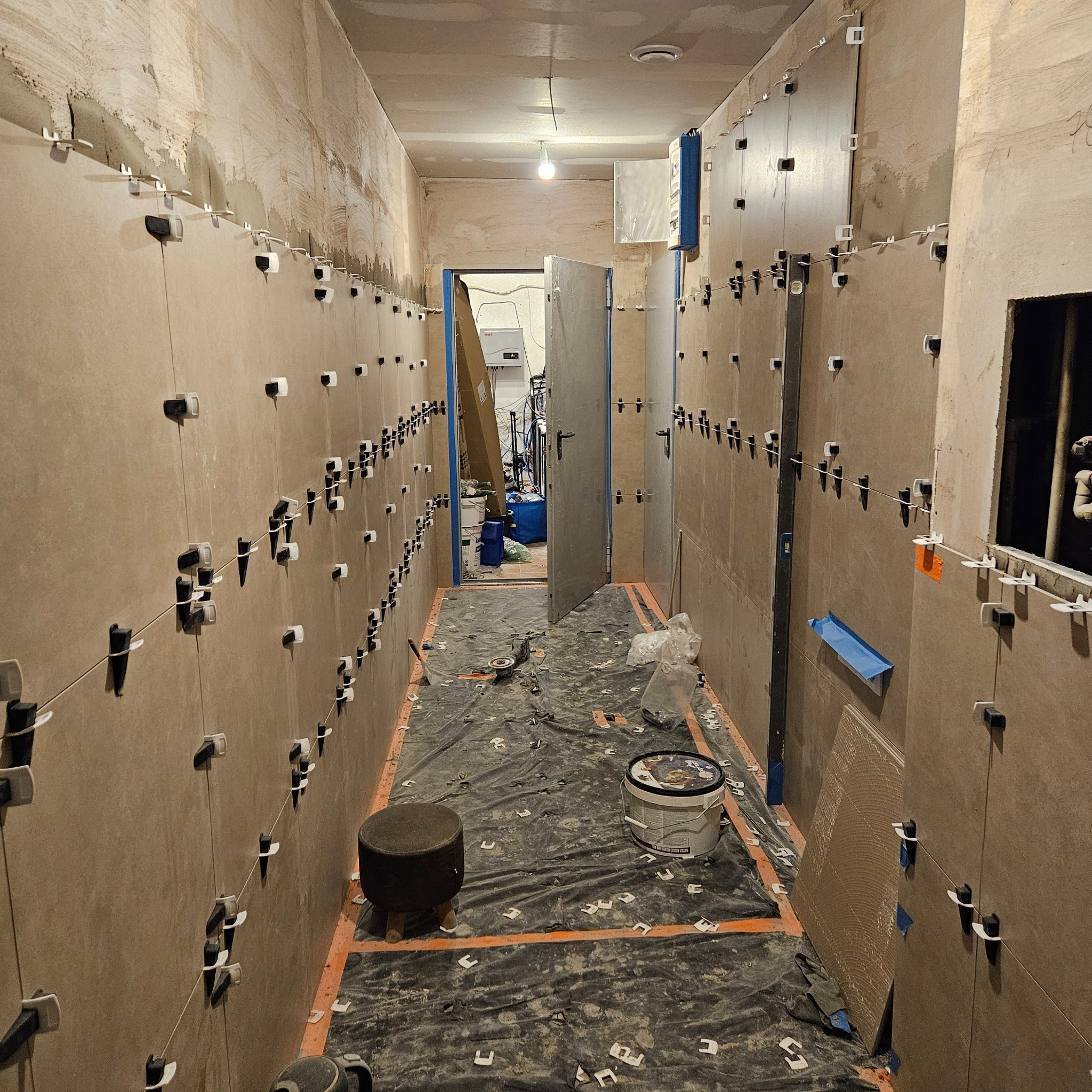 Korytarz w trakcie prac glazurniczych: beżowe płytki na ścianach, system poziomowania płytek, zabezpieczona podłoga. Widoczne narzędzia i materiały budowlane.