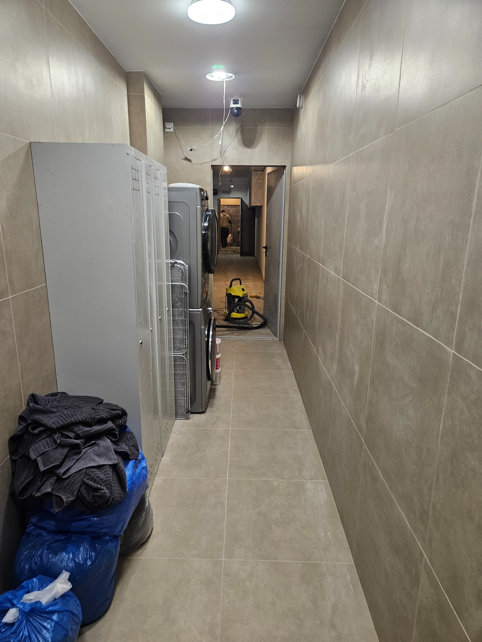 Wykafelkowane, wąskie pomieszczenie z pralkami i workami na podłodze. W tle widoczny odkurzacz i osoba w przejściu. Stonowana kolorystyka płytek.