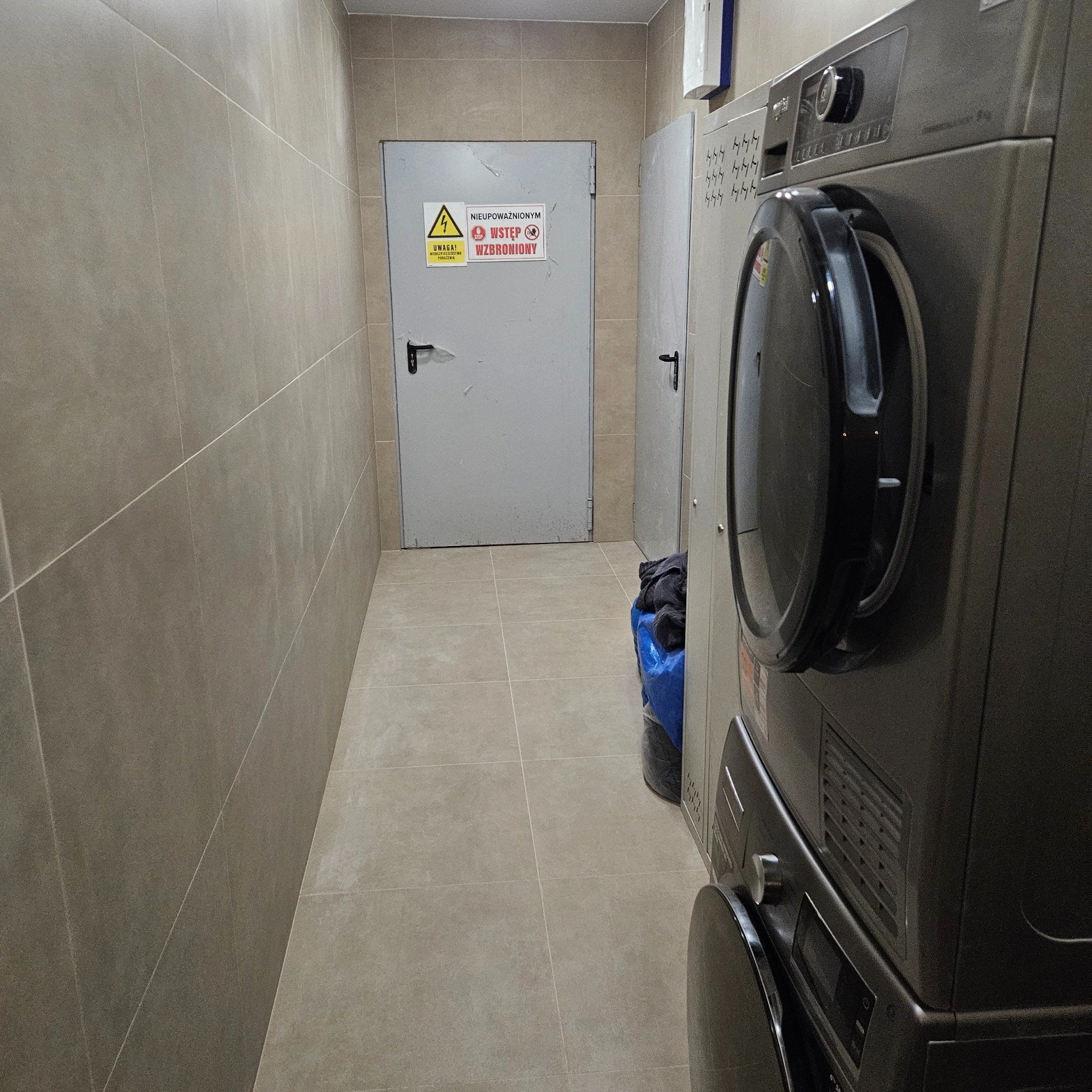 Wnętrze pomieszczenia z szarą pralką, wyłożonego beżowymi płytkami. Widoczne drzwi z ostrzeżeniem. Ujęcie z niskiego kąta, podkreślające przestrzeń i wykończenie.