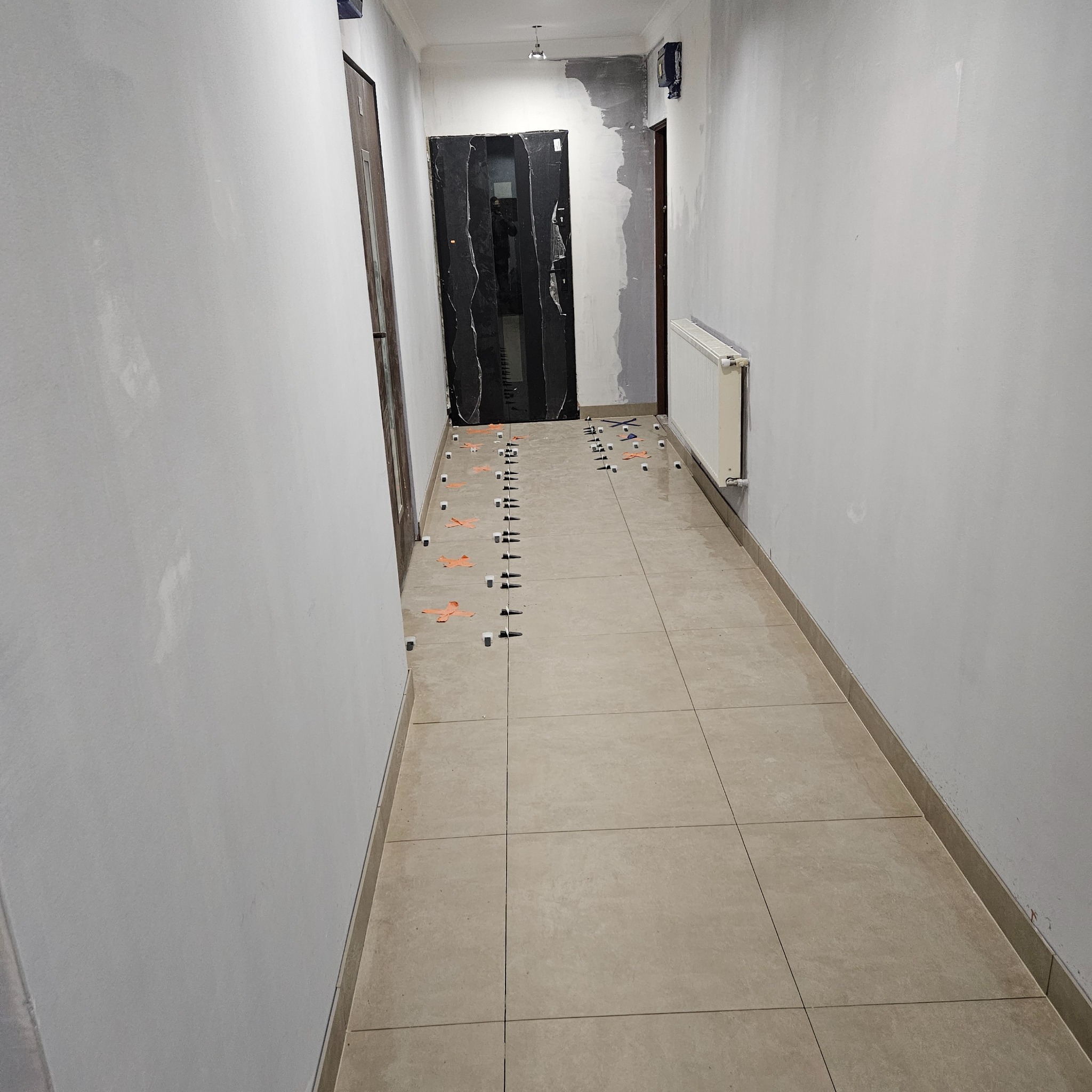 Korytarz w trakcie układania płytek podłogowych. Widoczne krzyżyki dystansowe i kliny poziomujące. Ściany przygotowane do malowania, grzejnik i drzwi po lewej stronie.