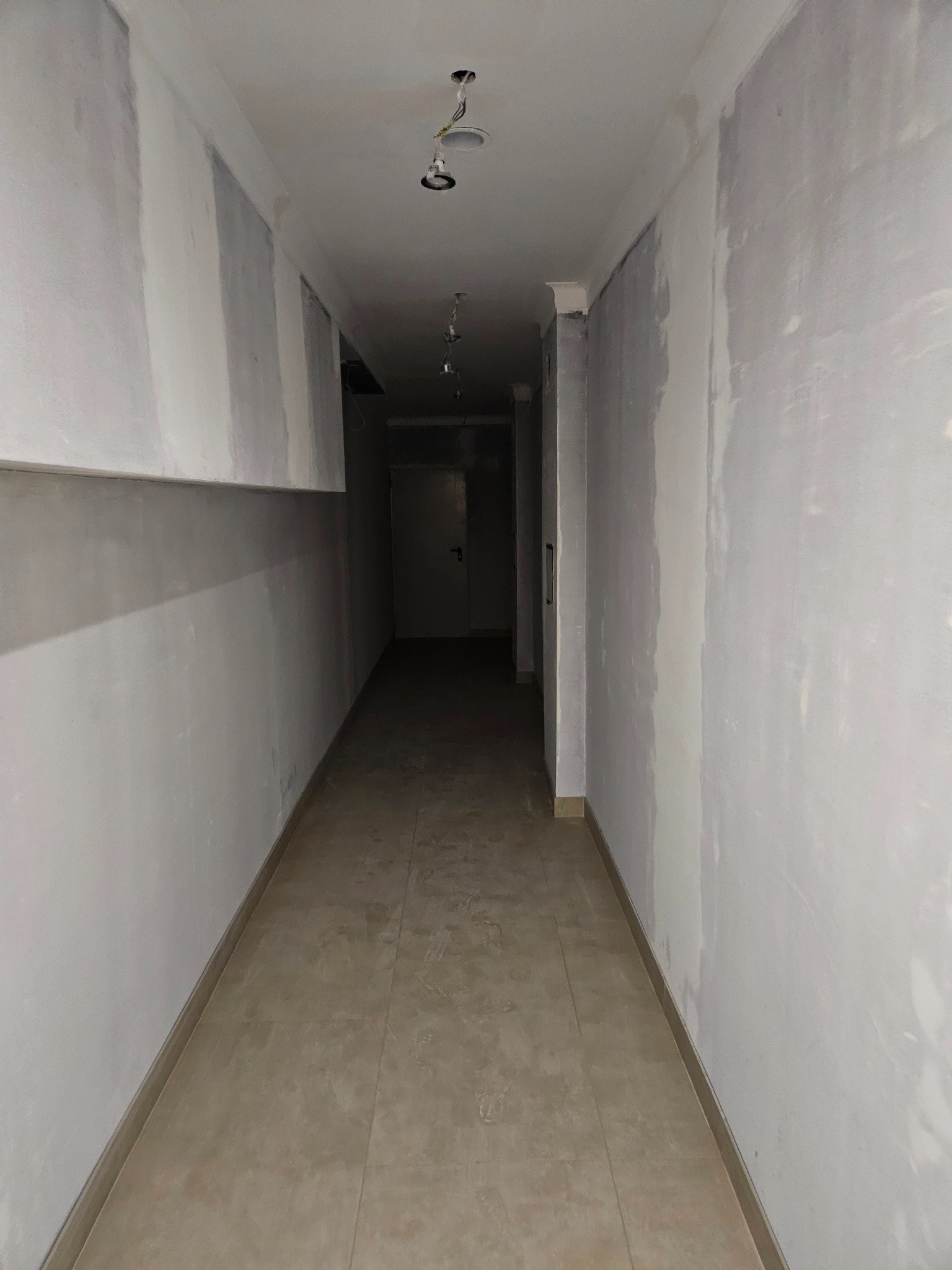 Korytarz w trakcie prac wykończeniowych: ściany po szpachlowaniu, widoczne punkty oświetleniowe na suficie i ułożona podłoga z płytek. Perspektywa z głębi korytarza.