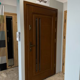 Vizera Sp. z o.o. - Brązowe drzwi wewnętrzne z przeszkleniami, prezentowane w salonie, z widocznym szyldem producenta 'Agmar' i fragmentami innych modeli drzwi w tle.