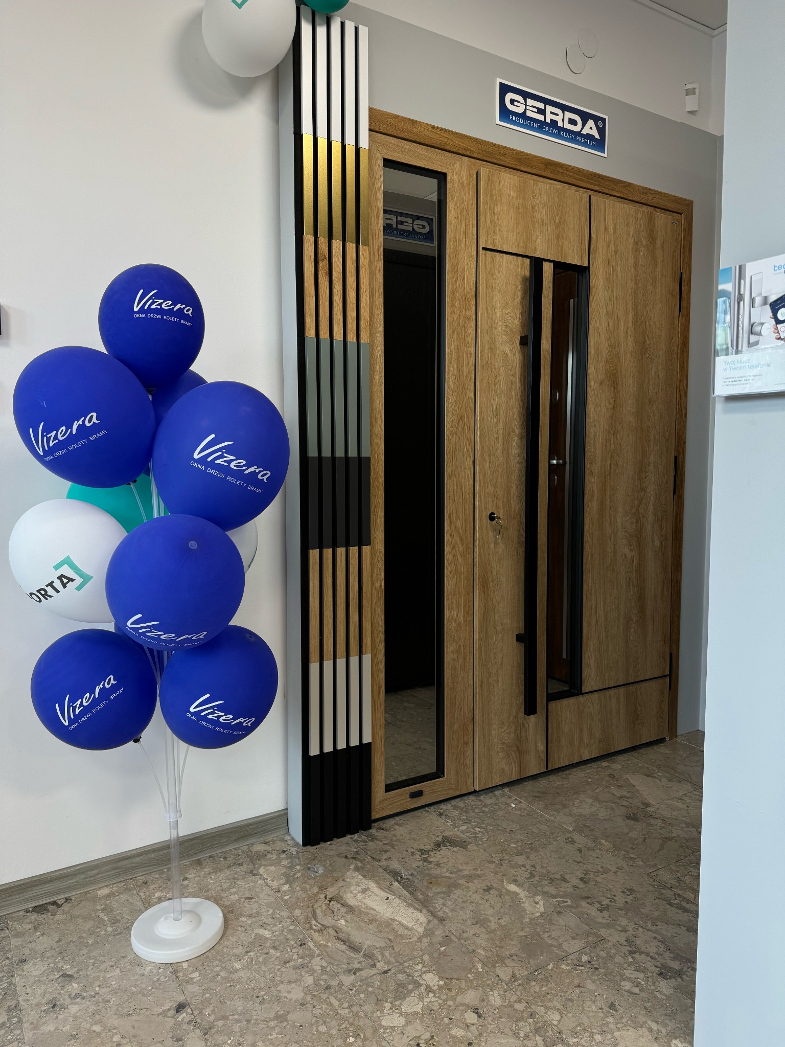 Wystawka drzwi Gerda z drewnianym wykończeniem i czarnymi elementami, obok balony z logo Vizera i Orta, prezentacja w salonie.