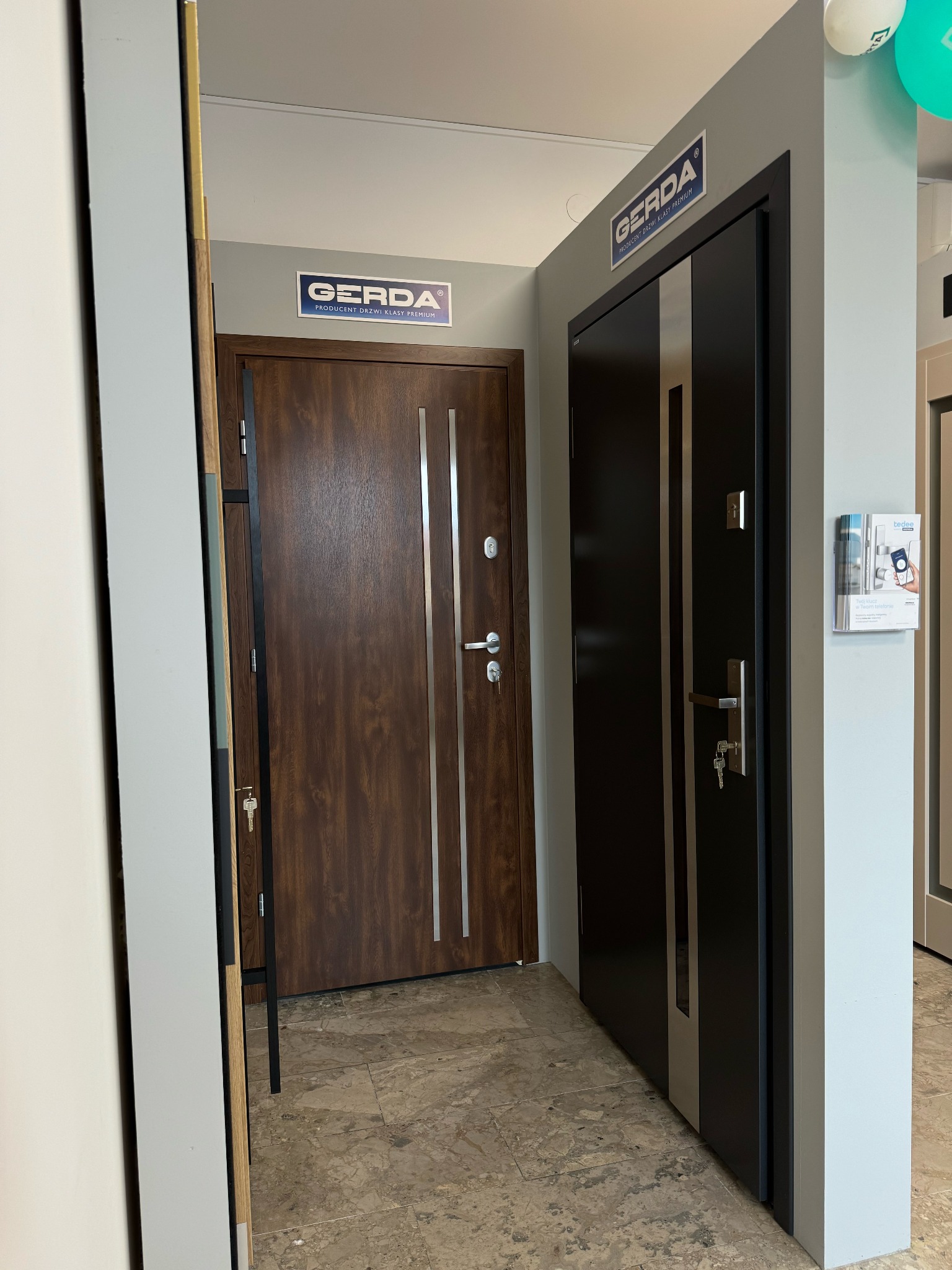 Ekspozycja dwóch modeli drzwi Gerda: brązowe drzwi z pionowymi, srebrnymi wstawkami i czarne drzwi z jedną pionową, szarą wstawką. Widoczne klamki i zamki.