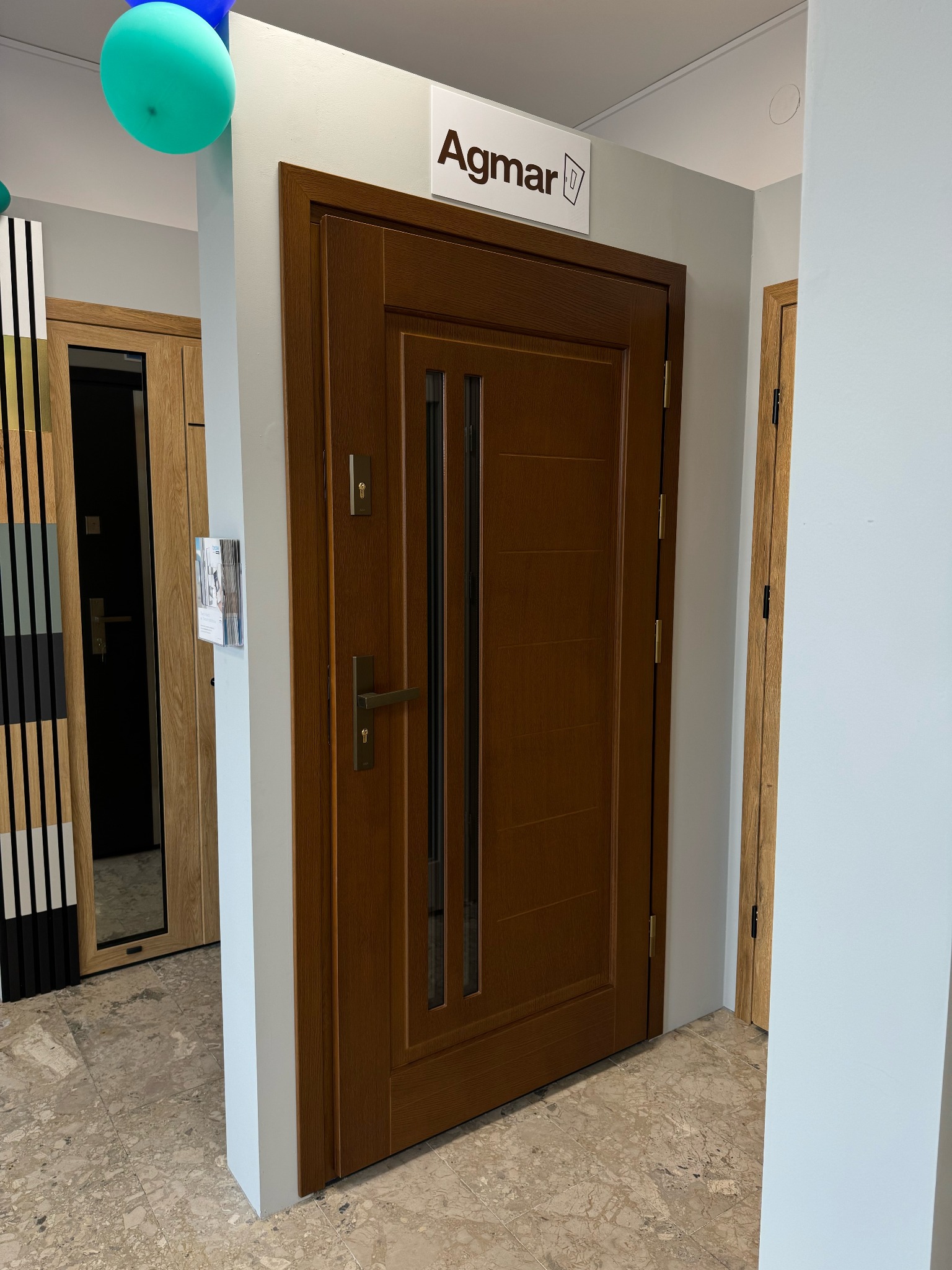 Brązowe drzwi wewnętrzne z przeszkleniami, prezentowane w salonie, z widocznym szyldem producenta 'Agmar' i fragmentami innych modeli drzwi w tle.
