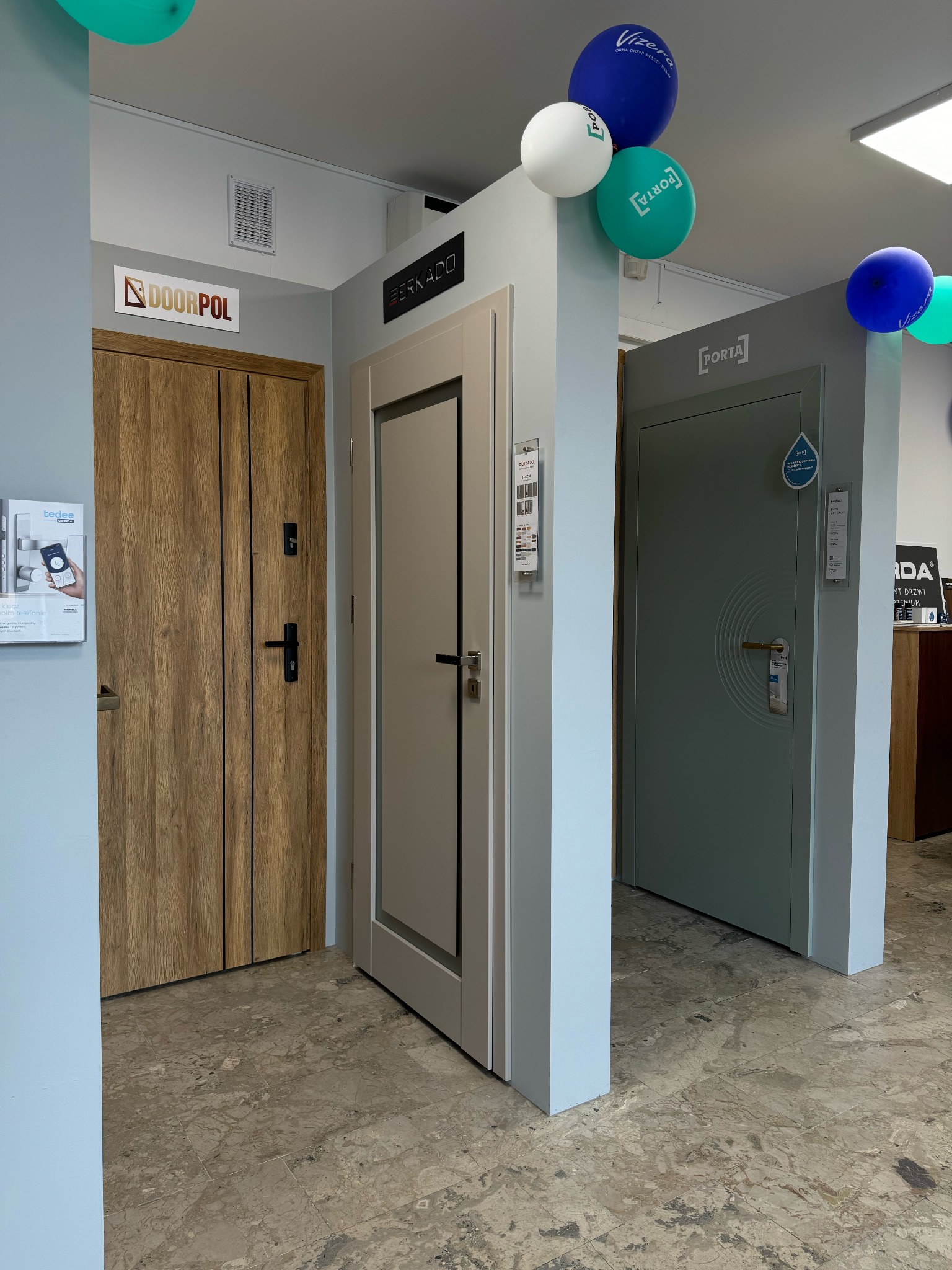 Ekspozycja trzech modeli drzwi wewnętrznych różnych producentów (Doorpol, Berkado, Porta) w salonie, z balonami reklamowymi pod sufitem i katalogami na ścianie.