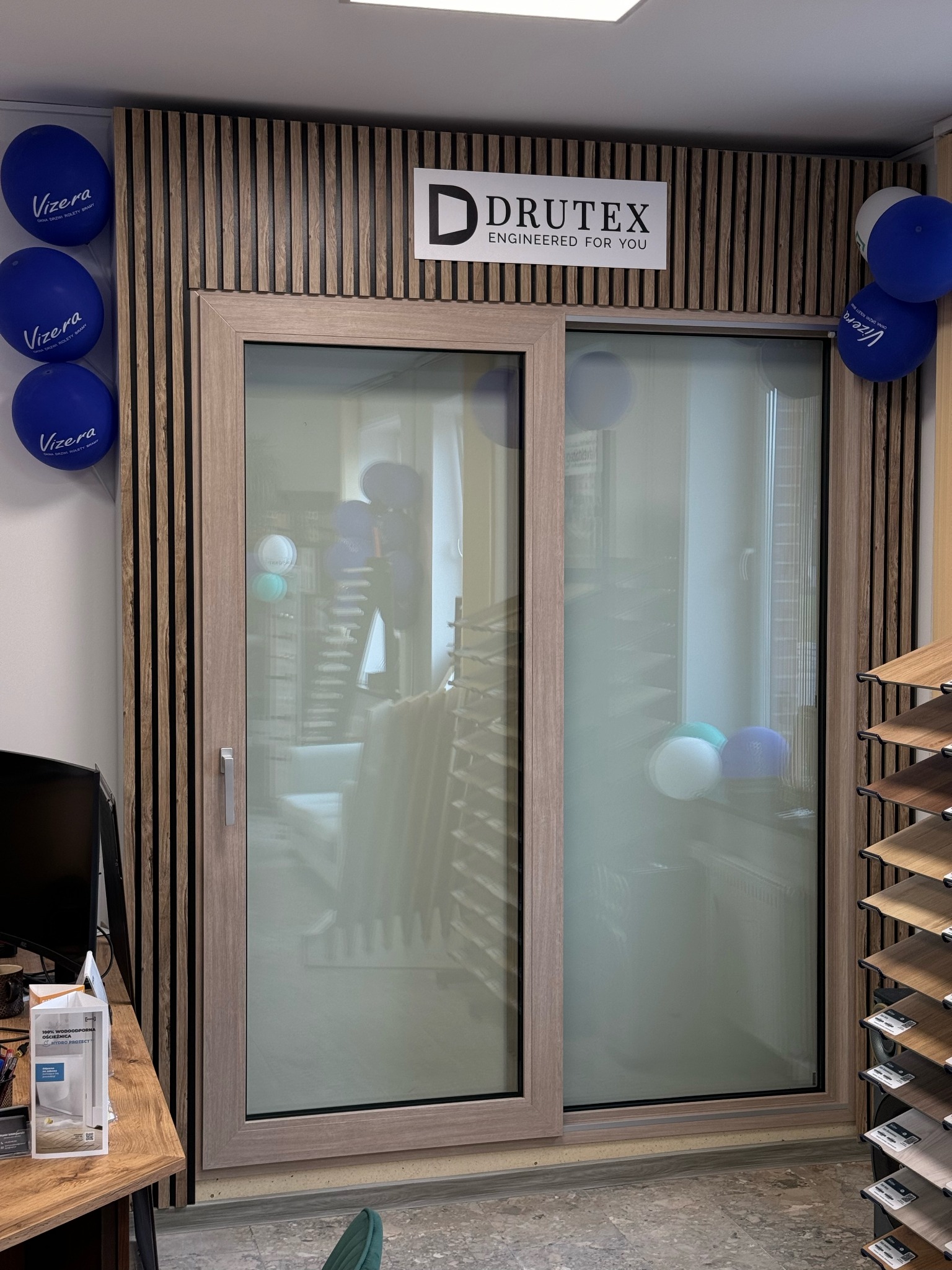 Przesuwne drzwi tarasowe Drutex z jasną ramą i szybami, z dekoracjami z balonów. W tle ekspozycja podłóg i ścianka z drewnianych listew.