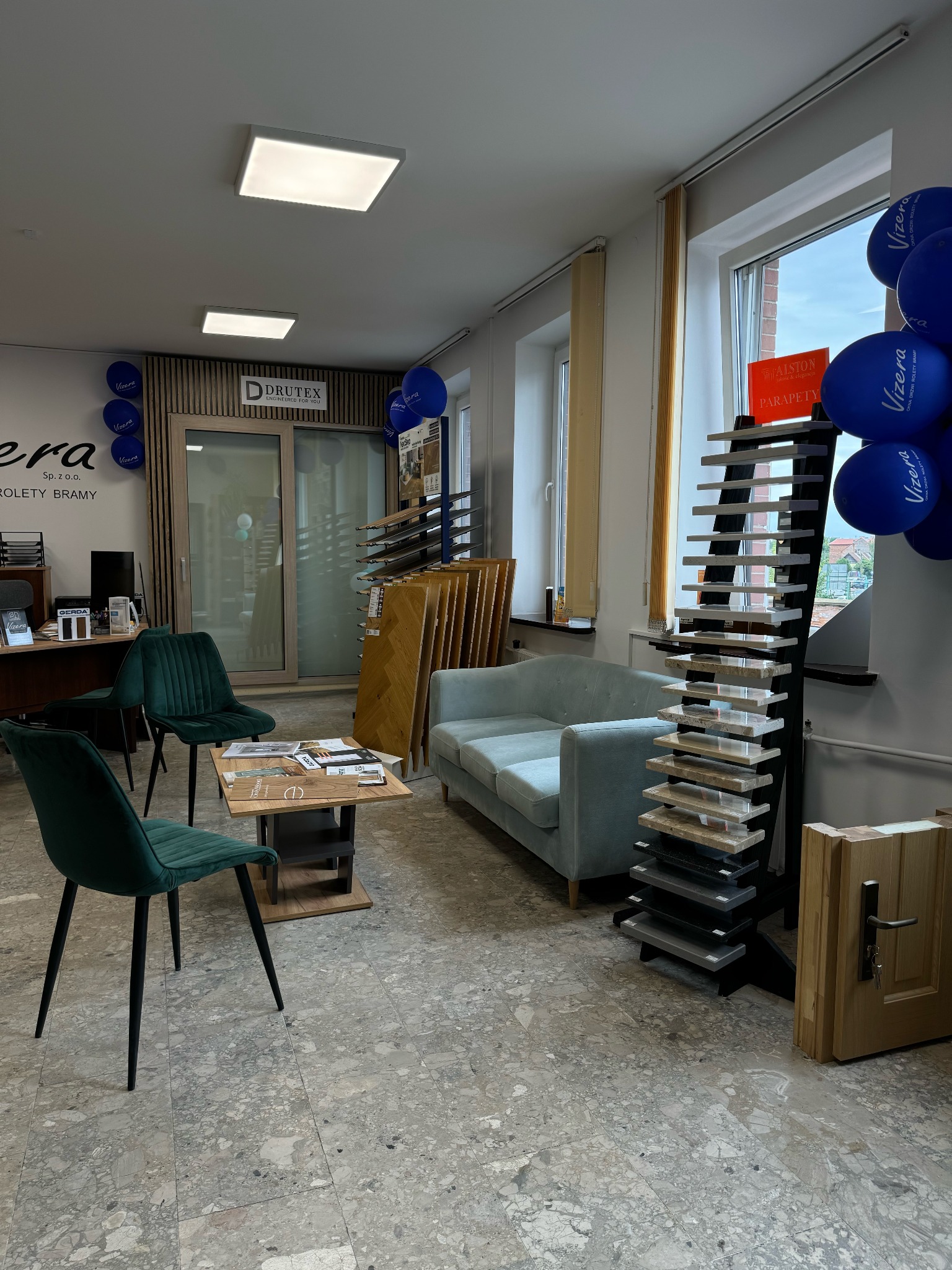 Salon z ekspozycją drzwi, podłóg i parapetów, z sofą i krzesłami dla klientów. Na oknach rolety, a na ścianie logo firmy VIZERA i DRUTEX.