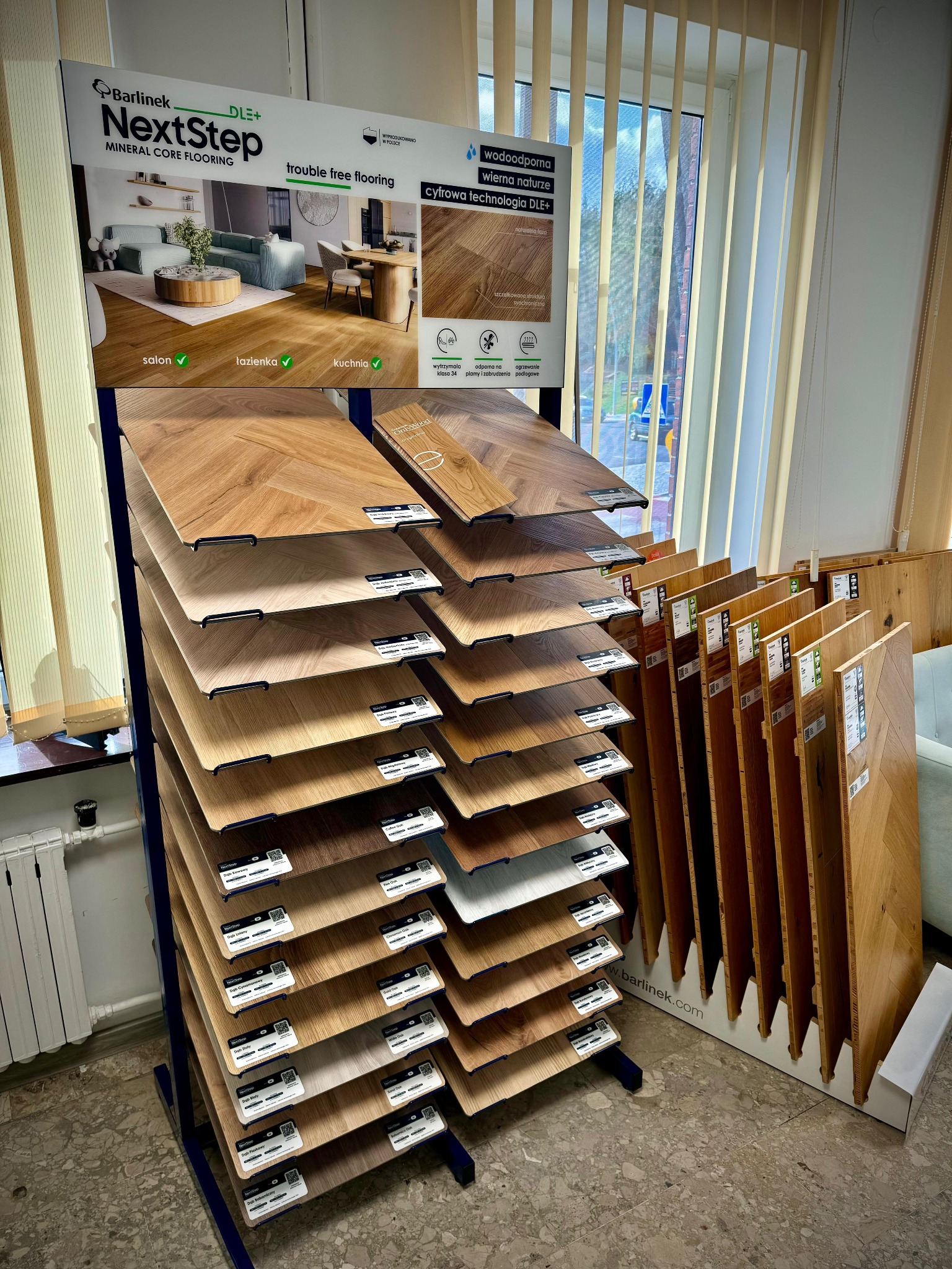 Ekspozycja paneli podłogowych Barlinek NextStep DLE+ na stojaku, prezentująca różne wzory i kolory, z oznaczeniami 'salon', 'łazienka', 'kuchnia'.