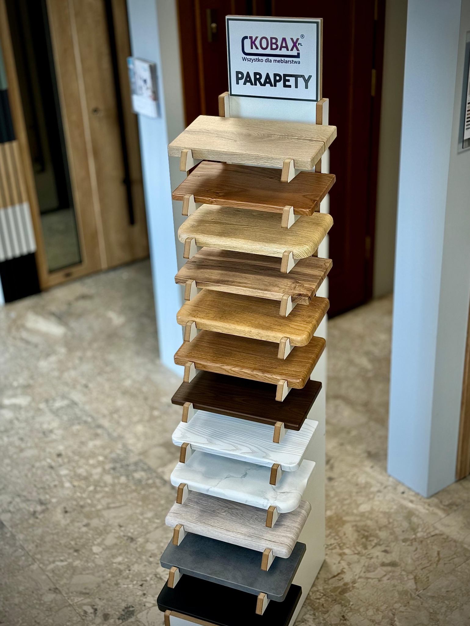 Ekspozycja próbek parapetów marki Kobax z różnymi wzorami i kolorami drewna oraz imitacji kamienia, prezentowana na stojaku w salonie meblowym.