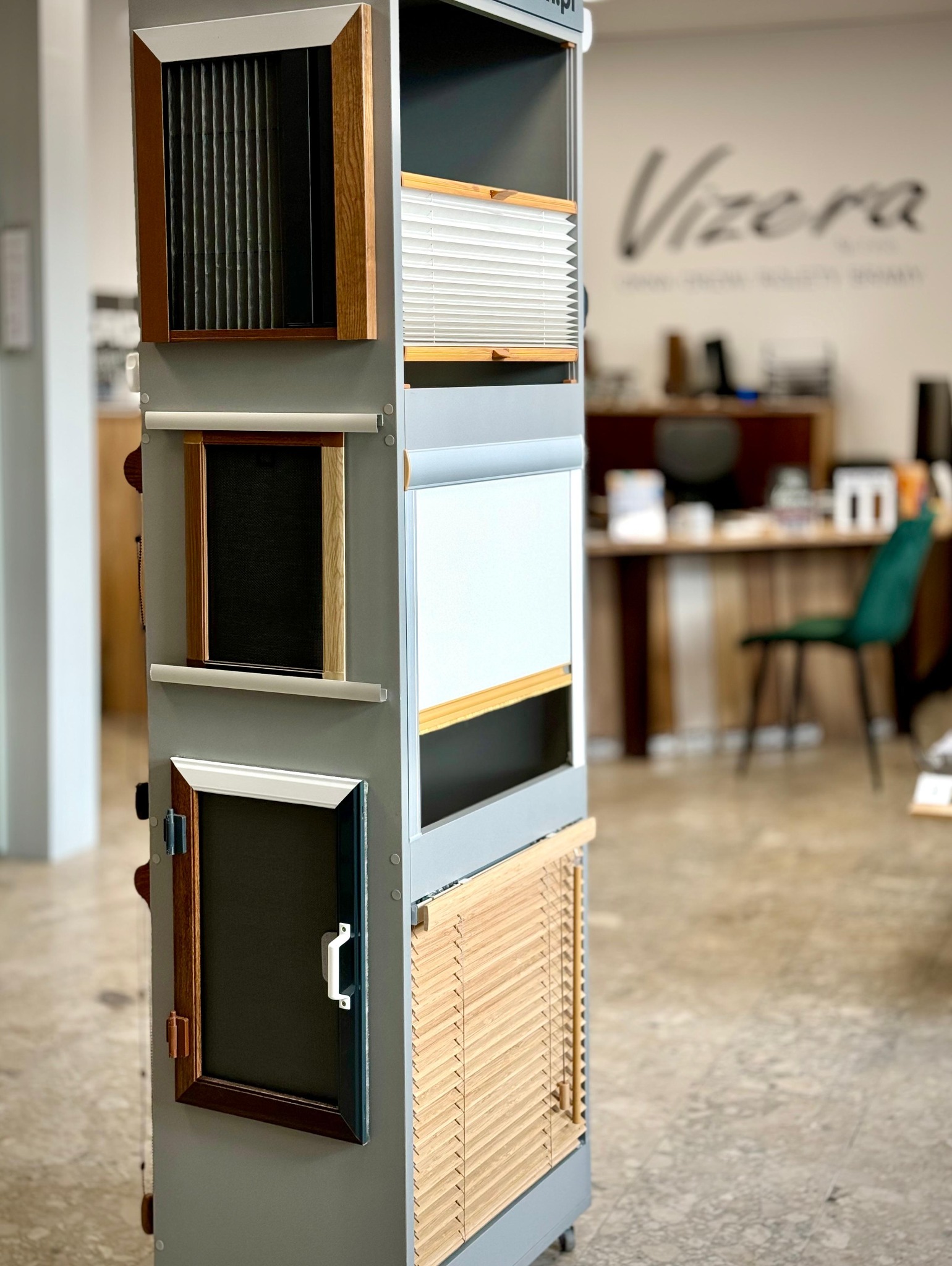 Ekspozytor z próbkami różnych typów osłon okiennych: plisy, żaluzje drewniane, rolety. Widoczne różne kolory i materiały. W tle logo firmy.