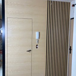 PPHU Mahoń stolarstwo Bogusław Nowak - Drewniana zabudowa ściany z ukrytymi drzwiami i lamelami w kolorze jasnego drewna. Widoczny domofon i fragment podłogi z czarnymi akcentami.