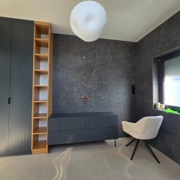 PPHU Mahoń stolarstwo Bogusław Nowak - Nowoczesna łazienka z szafką pod umywalkę w kolorze grafitowym, drewnianym regałem i krzesłem na tle ściany z szarych płytek. Minimalistyczny design, oświetlenie sufitowe.