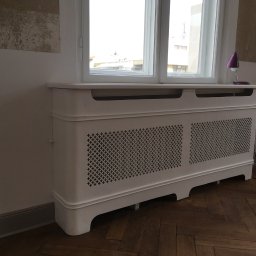 PPHU Mahoń stolarstwo Bogusław Nowak - Biała obudowa grzejnika z dekoracyjnym ażurem, ustawiona pod oknem. Na obudowie stoi fioletowa lampka. Parkiet w jodełkę na podłodze. Ściana z widocznymi śladami po remoncie.