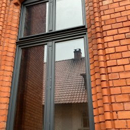 PPHU Mahoń stolarstwo Bogusław Nowak - Szare okno drewniane w łukowatej, ceglanej wnęce. W szybach odbija się budynek z ceglanym dachem i kominem. Dekoracyjne wykończenie ceglanej ściany.