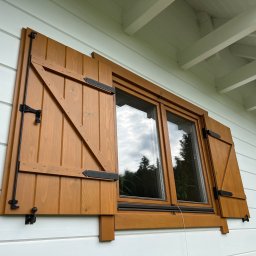 PPHU Mahoń stolarstwo Bogusław Nowak - Drewniane okno z otwartymi okiennicami w rustykalnym stylu, widoczne czarne okucia. Biała elewacja domu z drewnianą podbitką dachową.