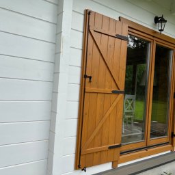 PPHU Mahoń stolarstwo Bogusław Nowak - Drewniane okiennice w kolorze miodowym z czarnymi okuciami, zamontowane na białej, drewnianej elewacji domu. Widoczne okno z ramą w tym samym kolorze co okiennice.