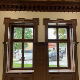 PPHU Mahoń stolarstwo Bogusław Nowak - Dwa drewniane okna z dekoracyjnymi ramami i zabudową grzejników, stylizowane na klasyczne, z widokiem na ulicę i zieleń. Górna część ściany zdobiona ornamentami.