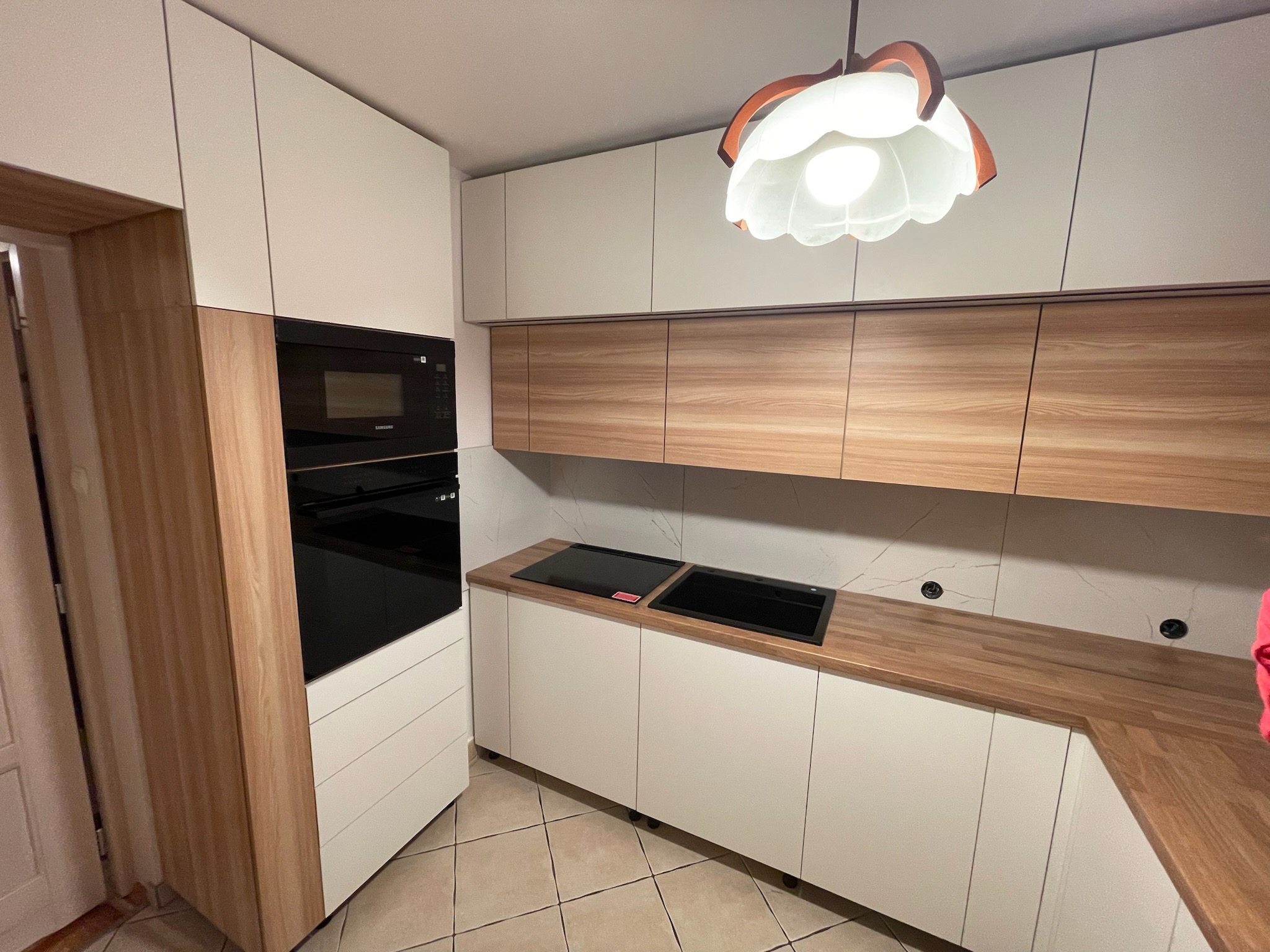 Nowoczesna kuchnia z białymi szafkami i drewnianymi blatami, oświetlona dekoracyjną lampą. Widoczny piekarnik i płyta grzewcza. Minimalistyczny design.