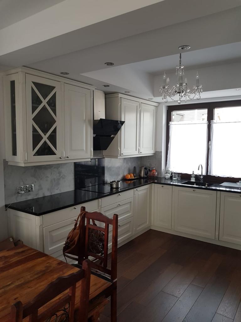 Jasna kuchnia w stylu klasycznym z czarnym blatem i drewnianą podłogą. Widoczny fragment stołu i krzesła. Nad oknem kryształowy żyrandol.