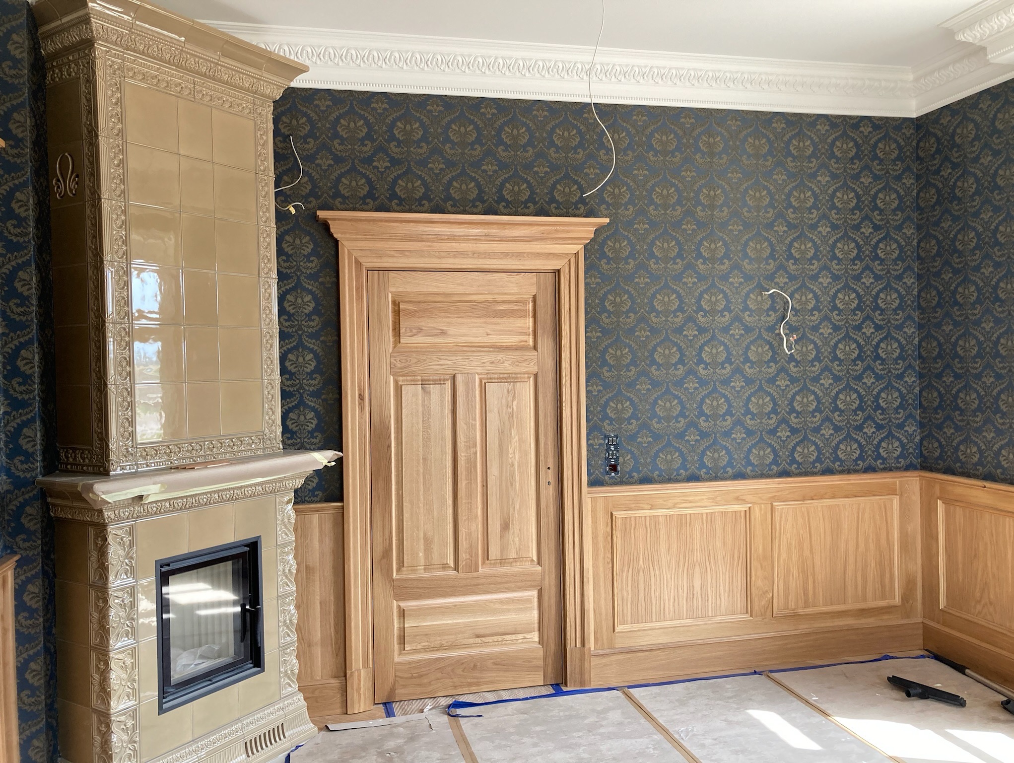 Klasyczne drzwi drewniane z płycinami i boazerią w odcieniu naturalnego drewna, na tle ściany w niebieskie wzory. Widoczny również piec kaflowy.