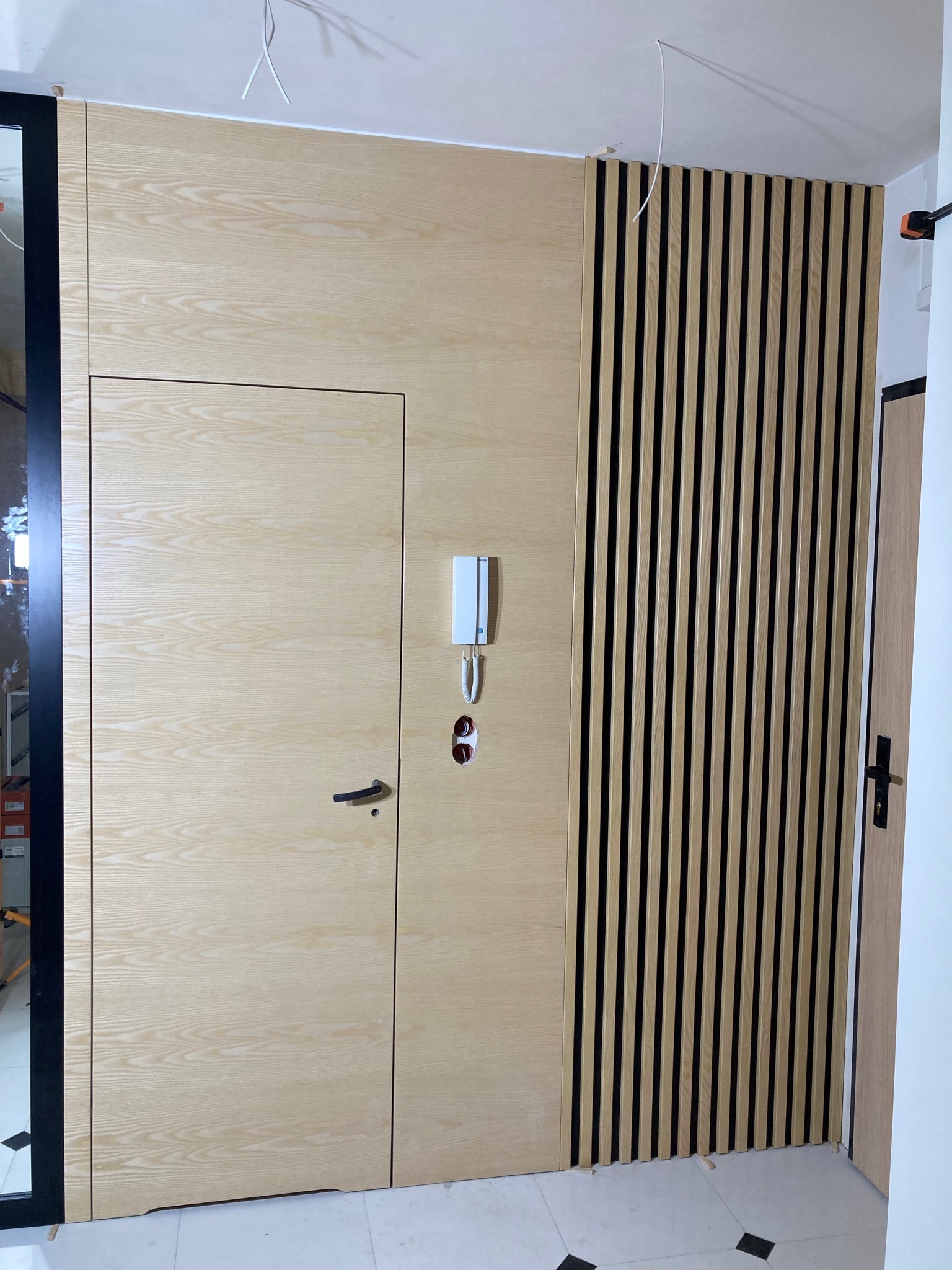 Drewniana zabudowa ściany z ukrytymi drzwiami i lamelami w kolorze jasnego drewna. Widoczny domofon i fragment podłogi z czarnymi akcentami.