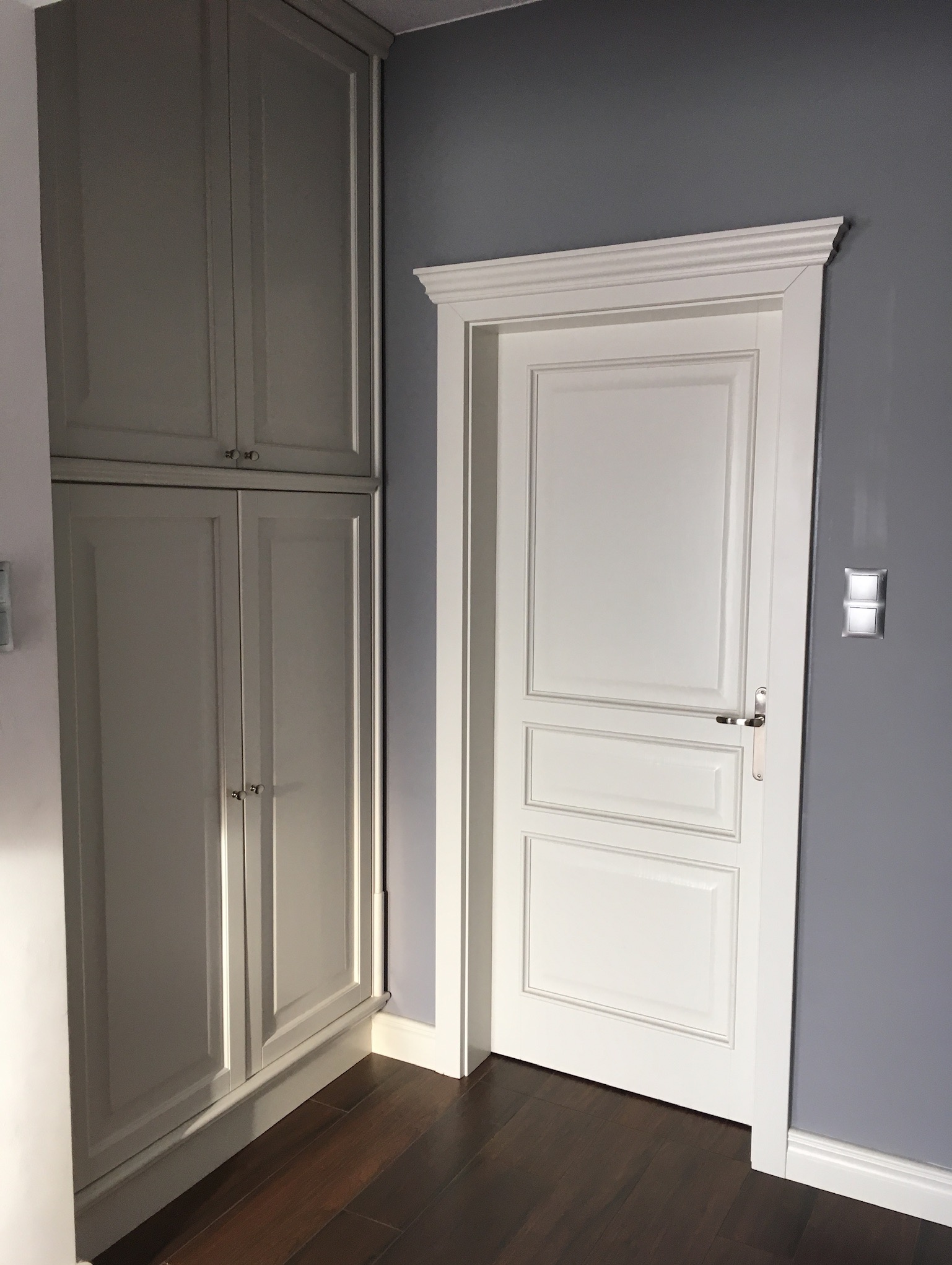 Białe, klasyczne drzwi wewnętrzne z ozdobną ramą, sąsiadujące z szarą szafą wnękową, na tle ściany w odcieniach szarości i ciemnej drewnianej podłogi.