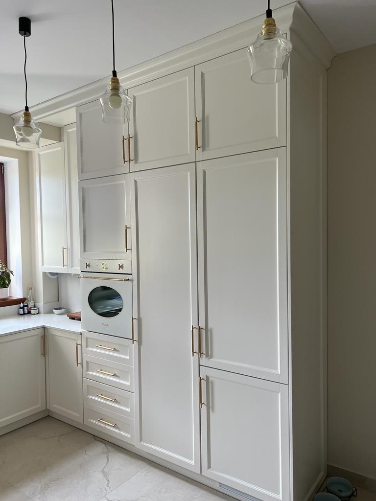 Elegancka, biała zabudowa kuchenna z piekarnikiem SMEG i złotymi uchwytami, oświetlona wiszącymi lampami w stylu retro. Klasyczny design z frezowanymi frontami.