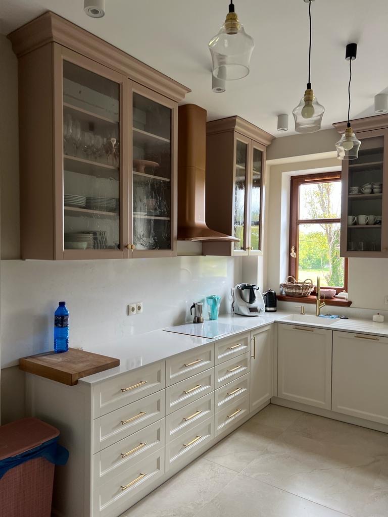 Jasna kuchnia z frezowanymi frontami, złotymi uchwytami i przeszklonymi szafkami wiszącymi. Nad blatem wisi miedziany okap. Nad wyspą kuchenną wiszą szklane lampy.
