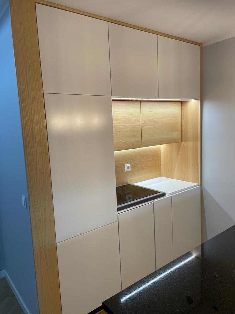 Nowoczesna zabudowa kuchenna z białymi frontami i drewnianymi akcentami, oświetleniem LED nad blatem i szafkami. Minimalistyczny design, widoczna płyta grzewcza.