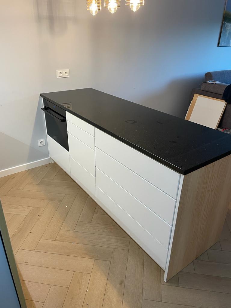 Wyspa kuchenna z białymi frontami i czarnym blatem, wykończona drewnem, na parkiecie ułożonym w jodełkę. Nad nią widoczna nowoczesna lampa.