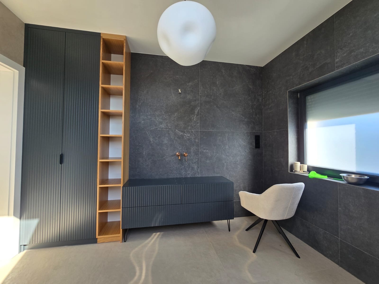 Nowoczesna łazienka z szafką pod umywalkę w kolorze grafitowym, drewnianym regałem i krzesłem na tle ściany z szarych płytek. Minimalistyczny design, oświetlenie sufitowe.