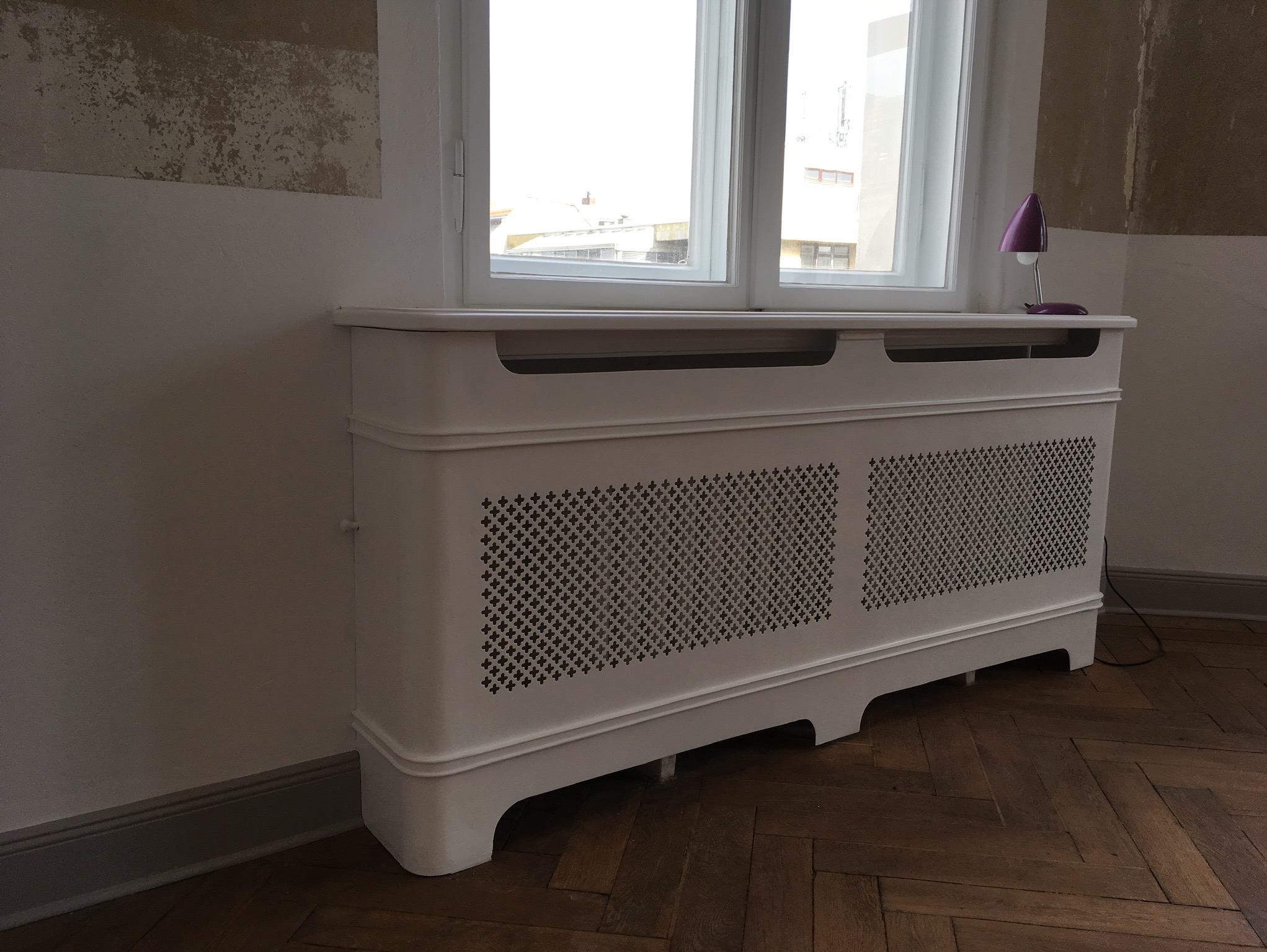 Biała obudowa grzejnika z dekoracyjnym ażurem, ustawiona pod oknem. Na obudowie stoi fioletowa lampka. Parkiet w jodełkę na podłodze. Ściana z widocznymi śladami po remoncie.