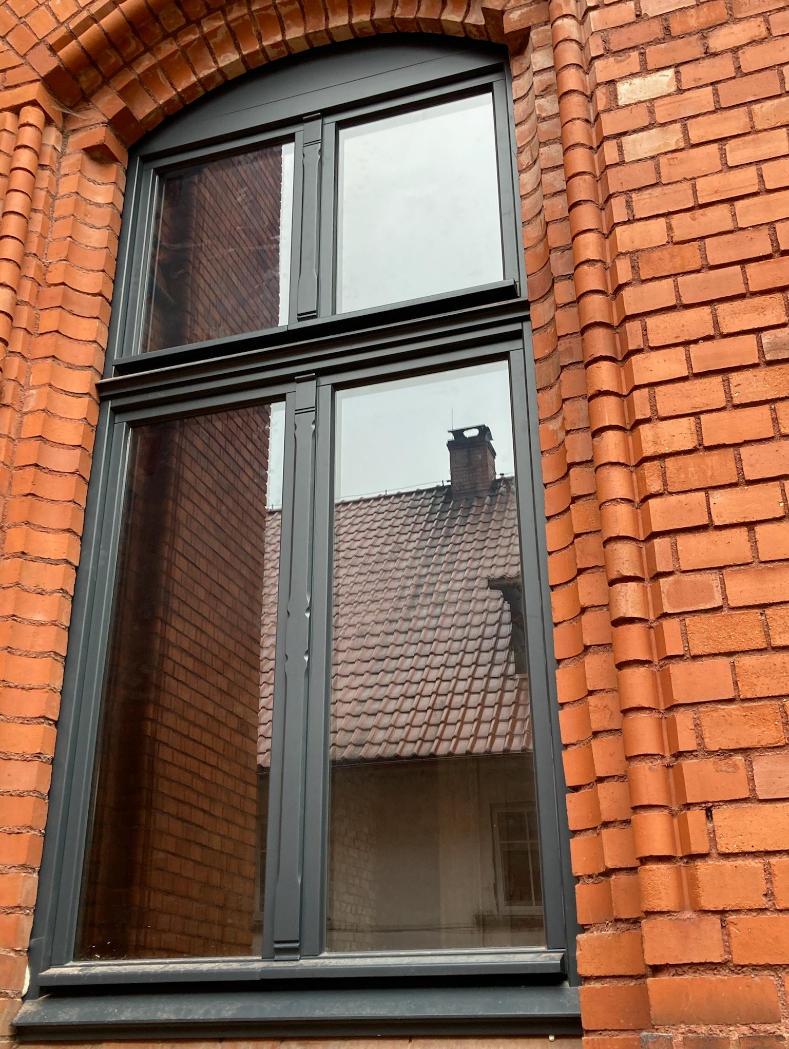 Szare okno drewniane w łukowatej, ceglanej wnęce. W szybach odbija się budynek z ceglanym dachem i kominem. Dekoracyjne wykończenie ceglanej ściany.