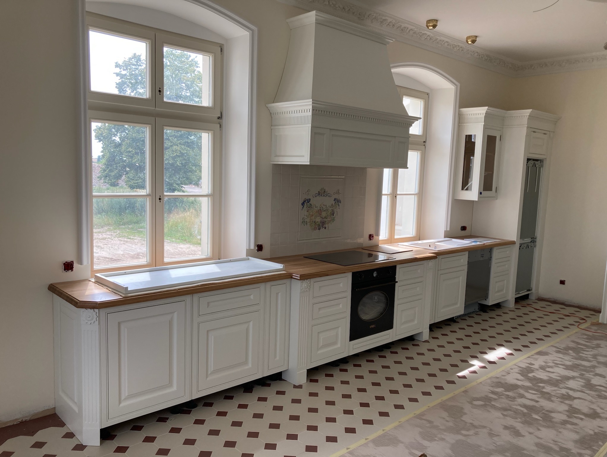 Jasna kuchnia w stylu klasycznym z drewnianym blatem, białymi frontami szafek z frezami i dużym oknem. Widoczny fragment podłogi z wzorem w romby.